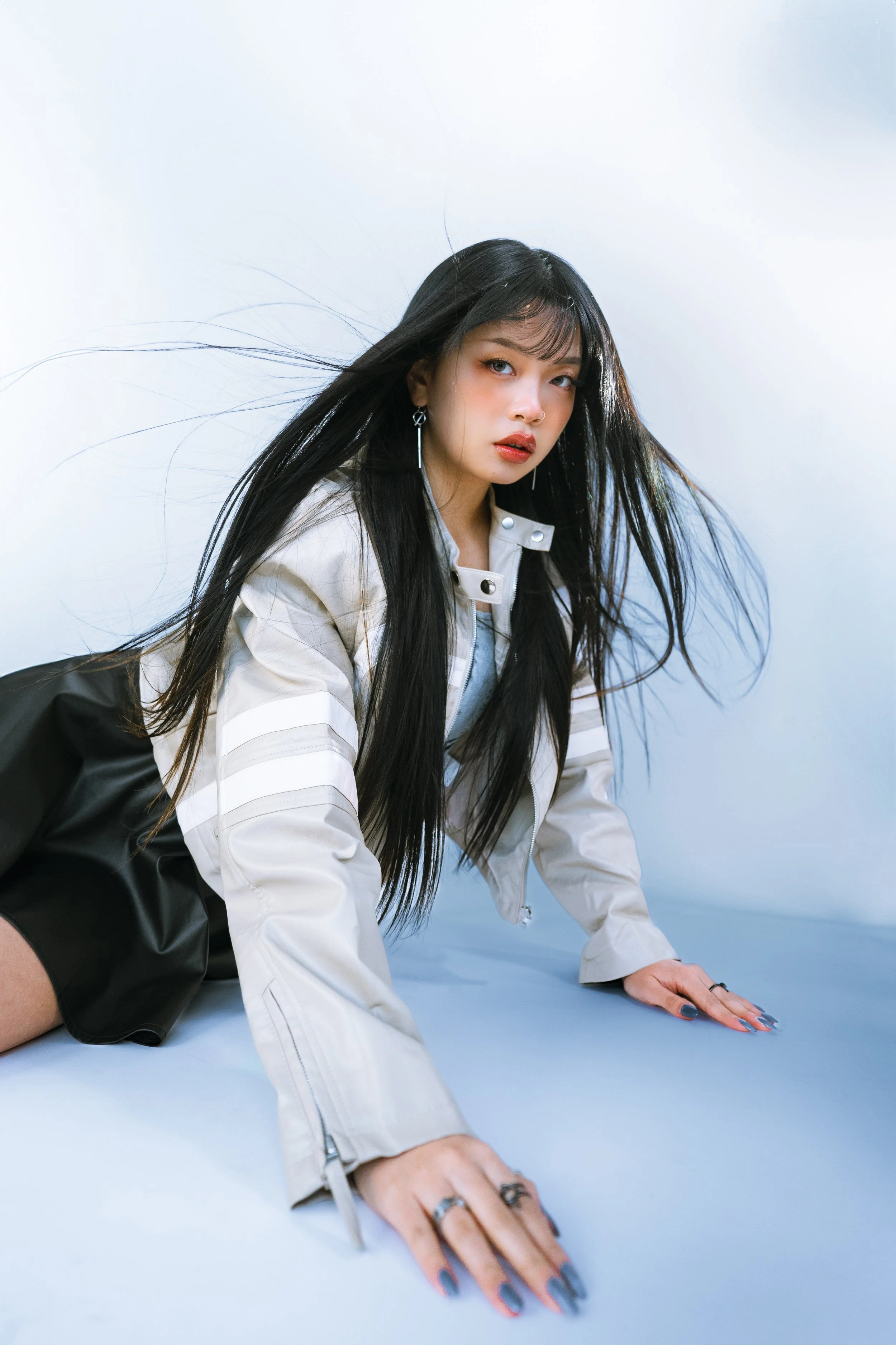 A young woman with long black hair and earrings lying on the ground with a blue background, wearing a white jacket and black shorts, looking at the camera.