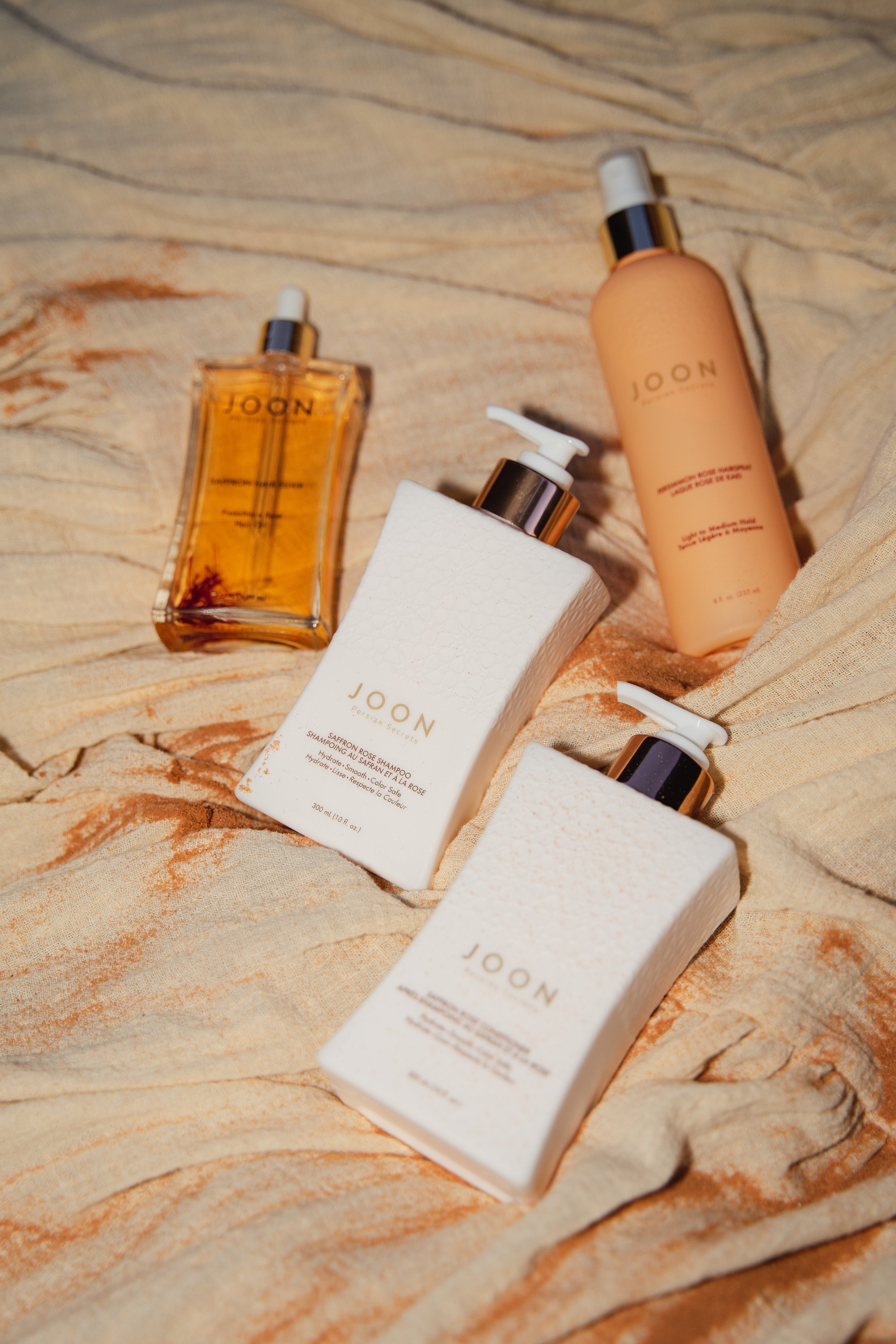 Collection of JOON skincare products, including two white bottles with pumps, one clear bottle with a dropper, and a beige spray bottle, arranged on a textured, beige fabric background.