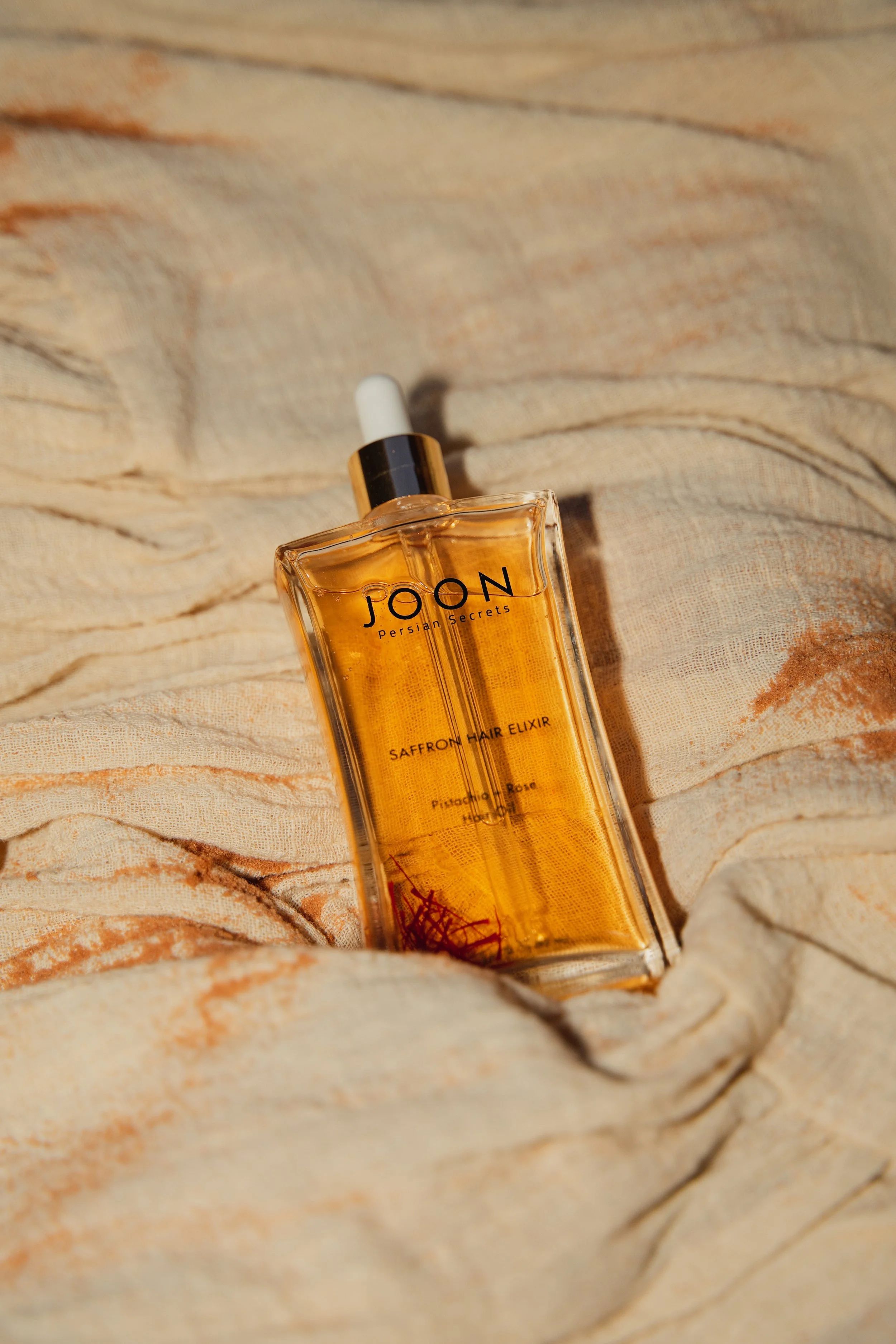 A bottle of JOON Persian Secrets Saffron Hair Elixir resting on light-colored, textured fabric.