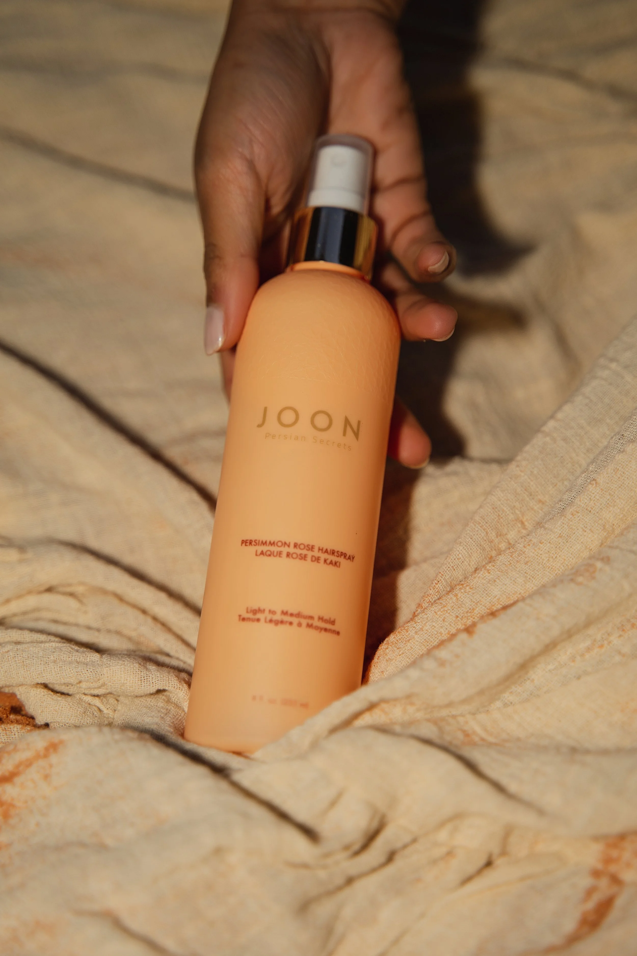 A hand holding a peach-colored bottle of JOON Persian Secrets Persemmom Rose Hairspray against a beige, textured fabric background.