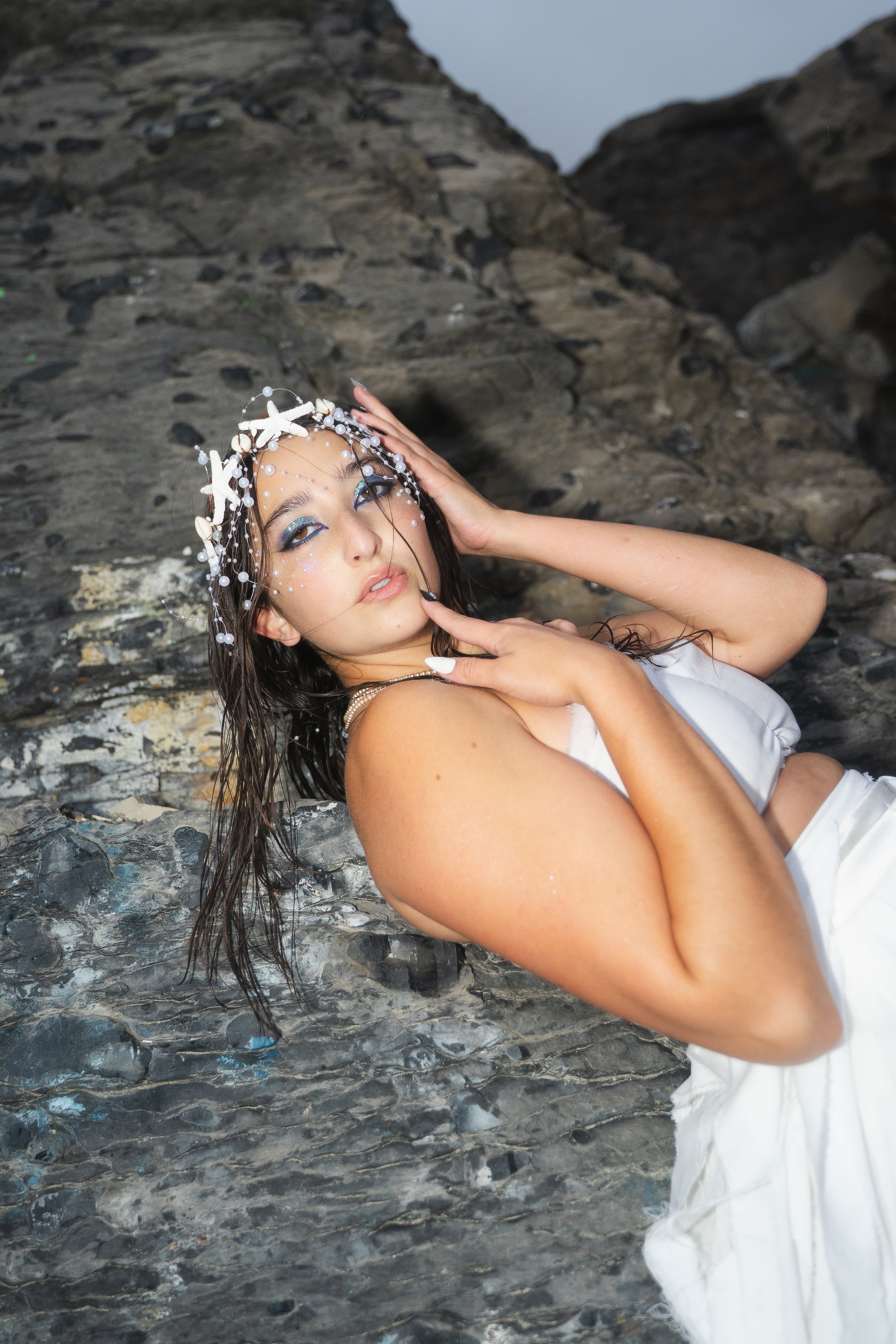 A woman with long dark damp hair, wearing a white dress and a headpiece decorated with white starfish and pearls, lying on a rocky beach with her hand on her head and looking at the camera.