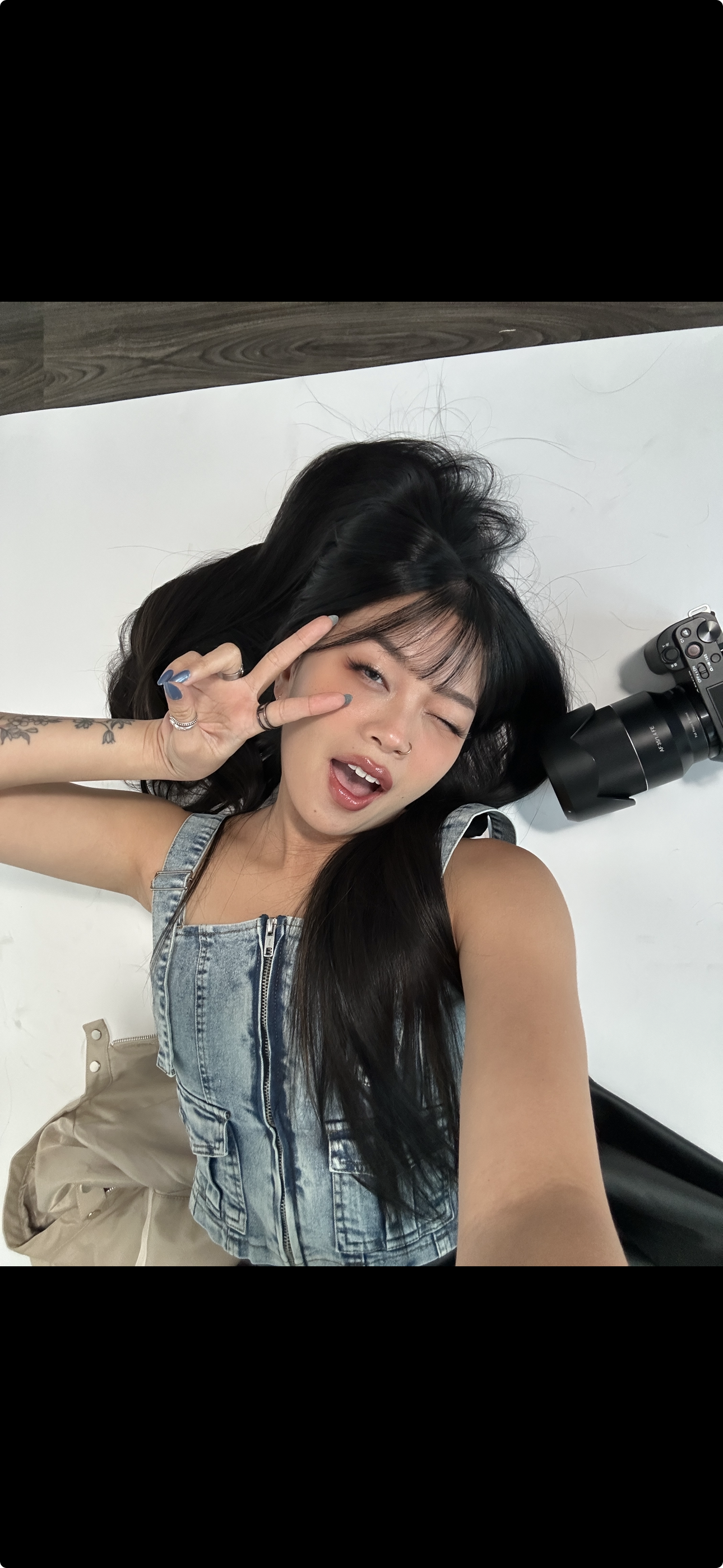 Young woman lying on a white surface, making a peace sign and winking, with a camera next to her.