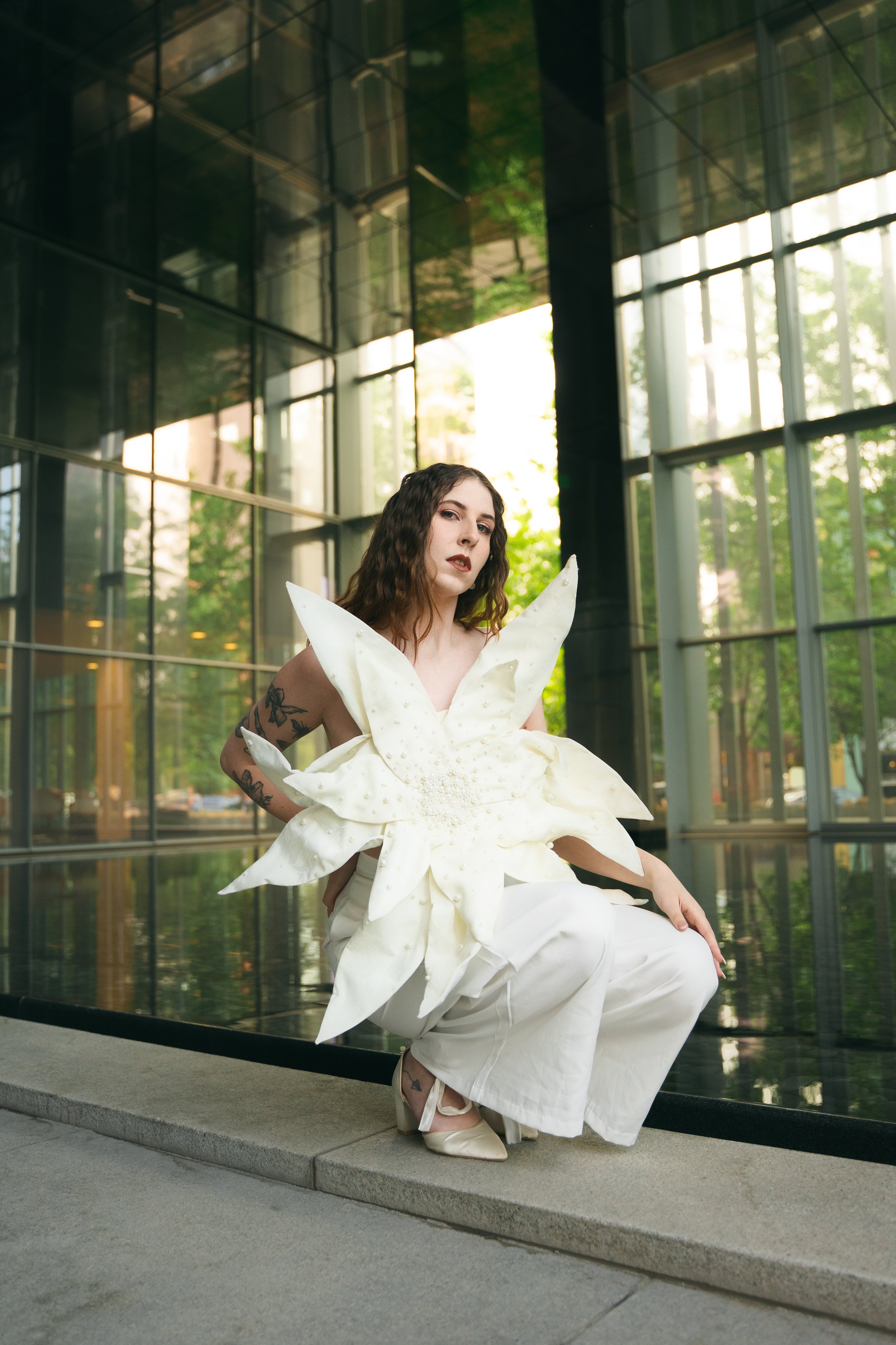 A woman with dark wavy hair, tattoos on her arm, and wearing a large white dress with petal-like features and pointed shoulders, is sitting on a low wall in front of glass windows with trees and sunlight reflecting in them.