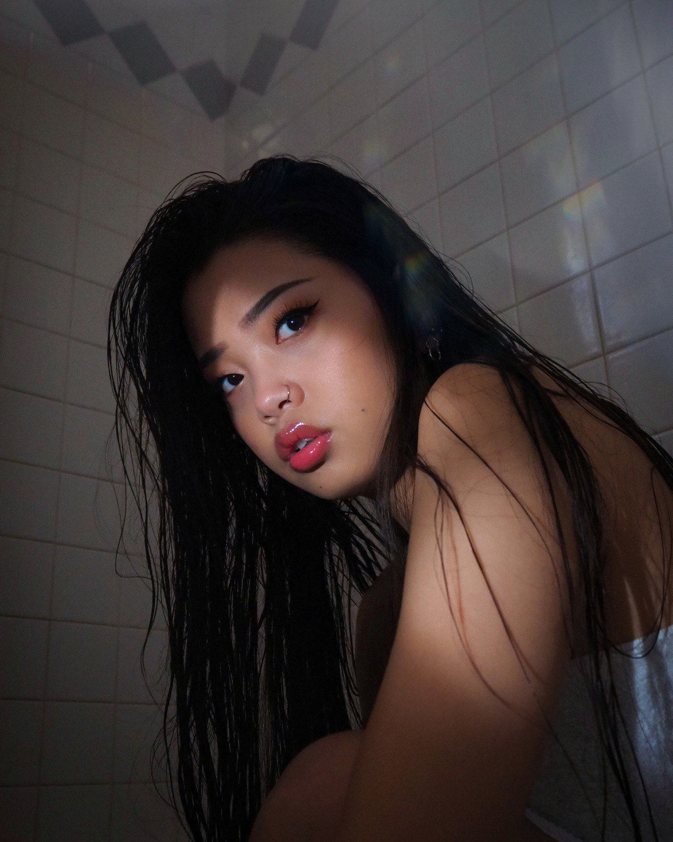 Close-up of a young woman with wet black hair, wearing makeup with winged eyeliner and glossy lips, in a tiled room, looking at the camera with a serious expression.