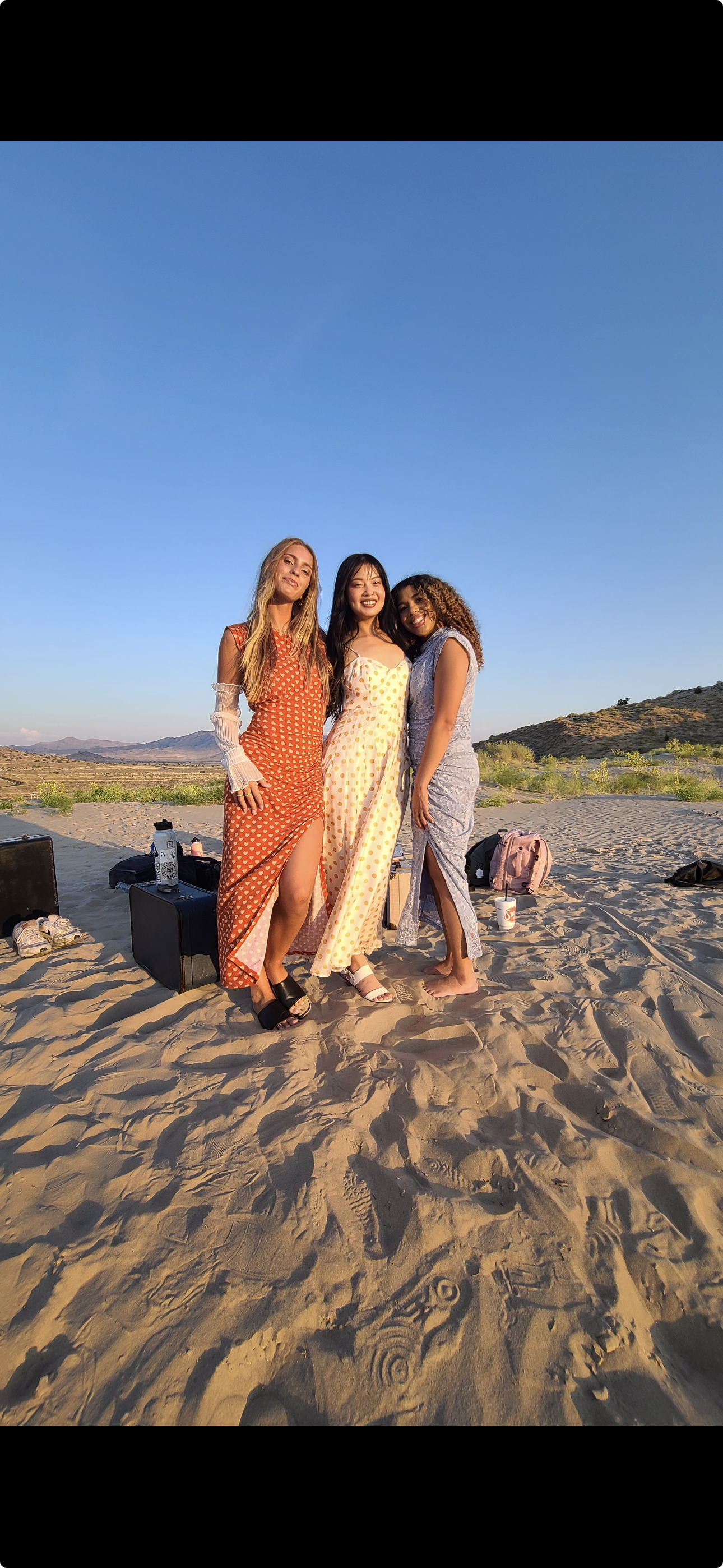 Three women standing together on a sandy desert with mountains and clear blue sky in the background.