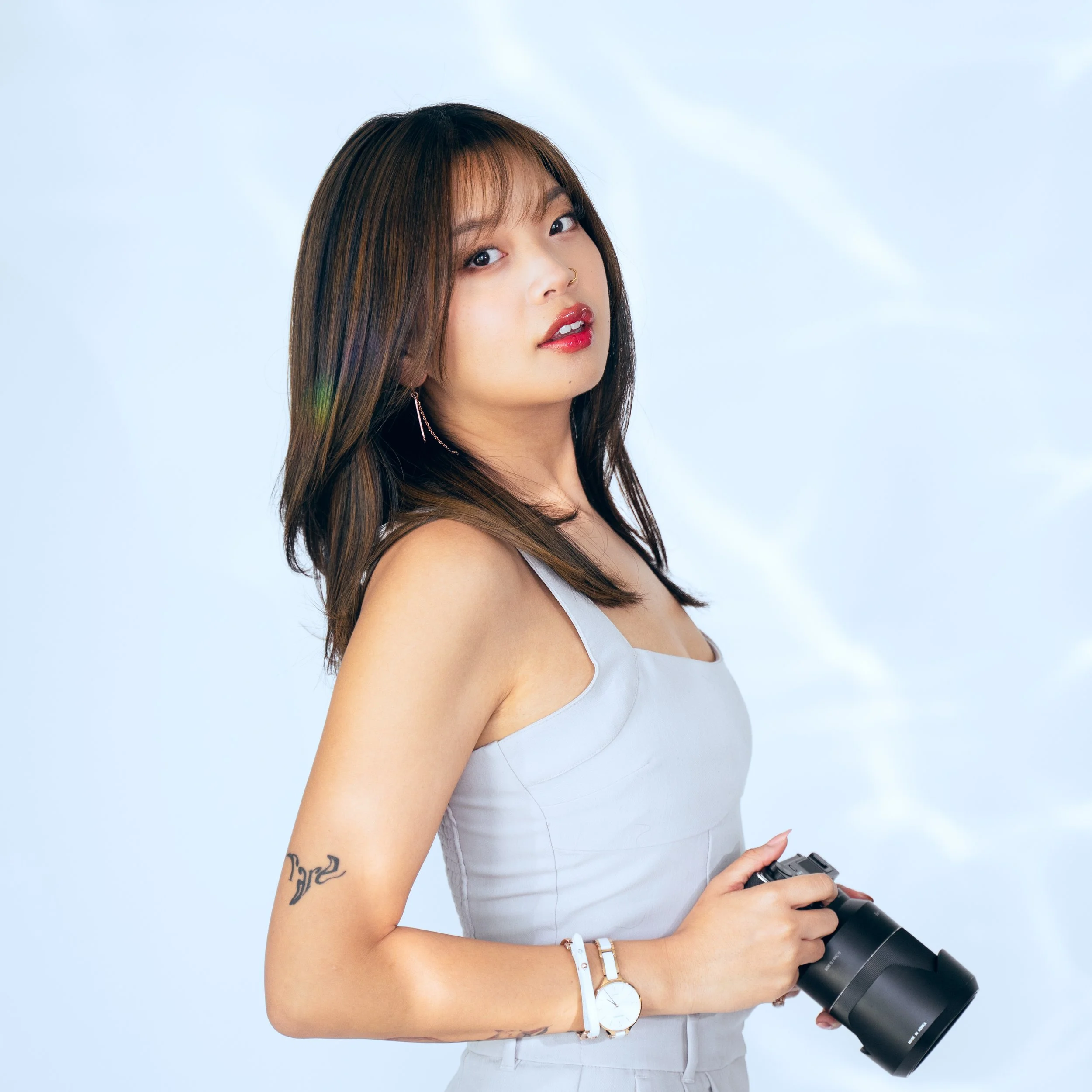 A young woman with shoulder-length brown hair holding a camera, wearing a sleeveless light-colored top, a white watch, and an earring, against a light blue background.
