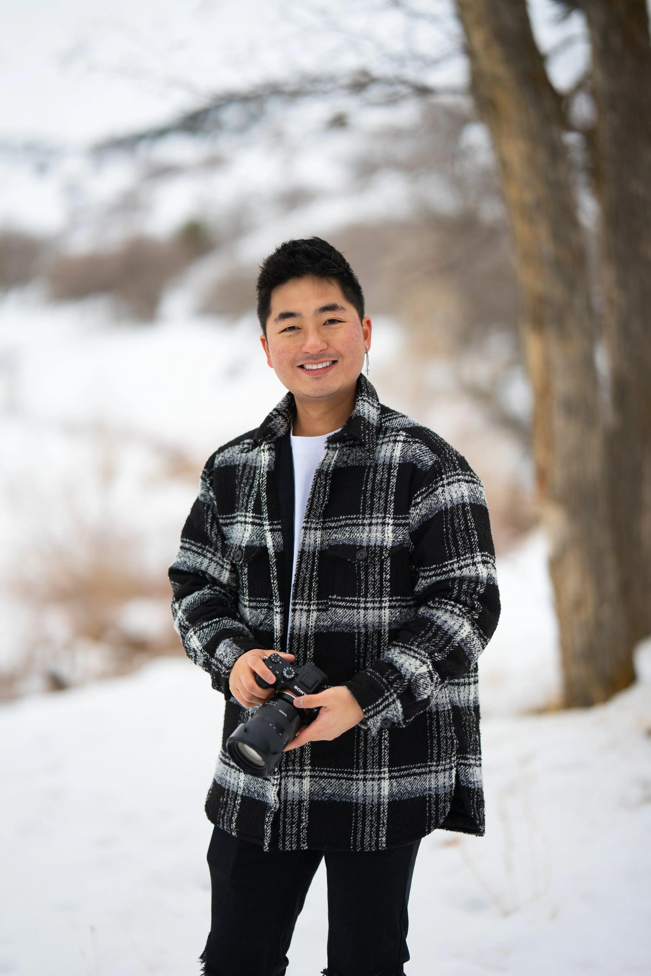 A young man smiling while holding a camera outdoors in a snowy landscape with trees in the background.