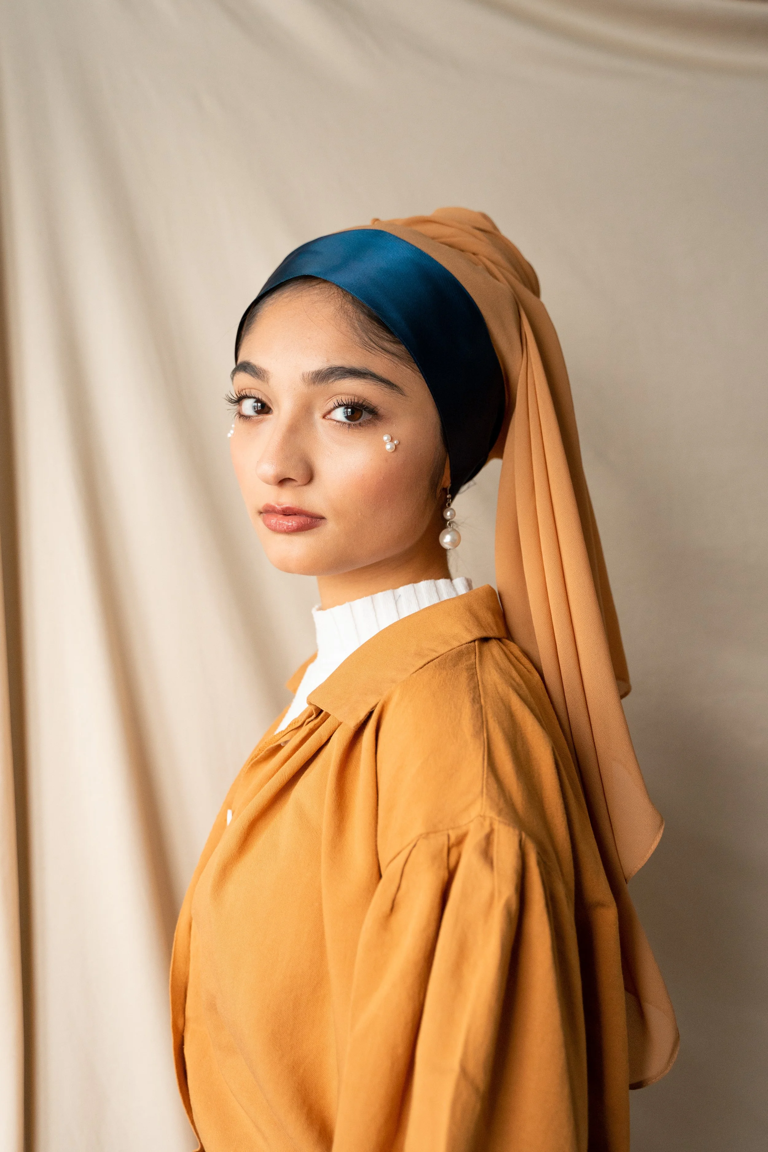 A woman with tan and dark blue headscarf, white top, and pearl jewelry, posing in front of a beige curtain background.