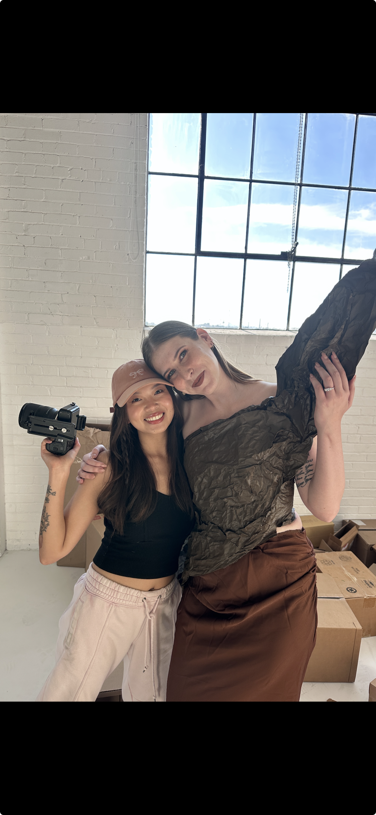 Two smiling women in casual clothing embrace inside a room with large windows and white brick walls. One woman holds a camera, and cardboard boxes are visible in the background.