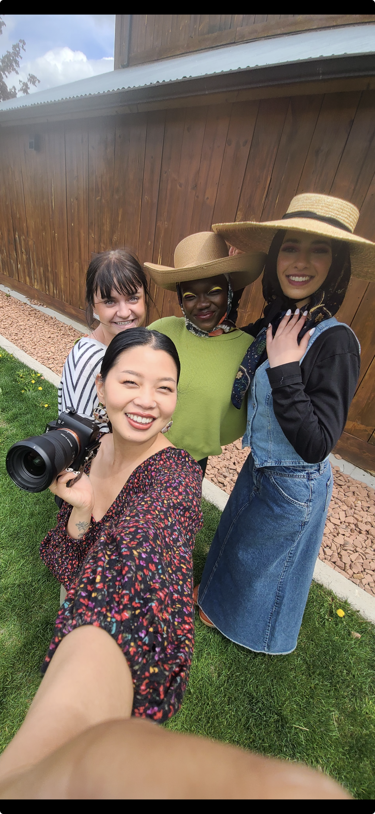 Group of four women outdoor taking selfie, smiling, with a wooden fence and green grass in background, two wearing large straw hats, one holding a camera.