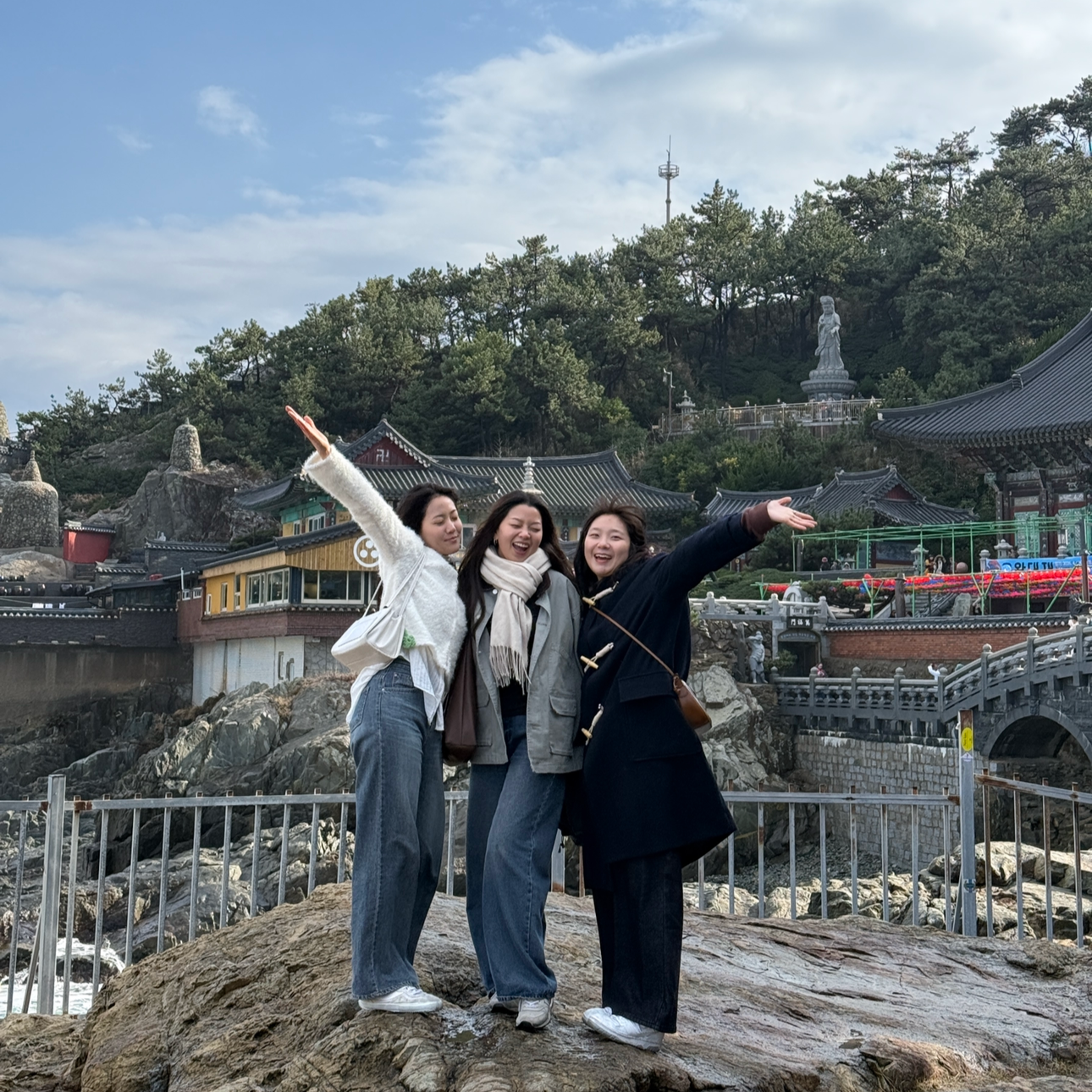 Three women standing on rocks near the water, smiling and posing with arms outstretched, in front of traditional Asian buildings and a hillside with a large statue of a woman in the background.