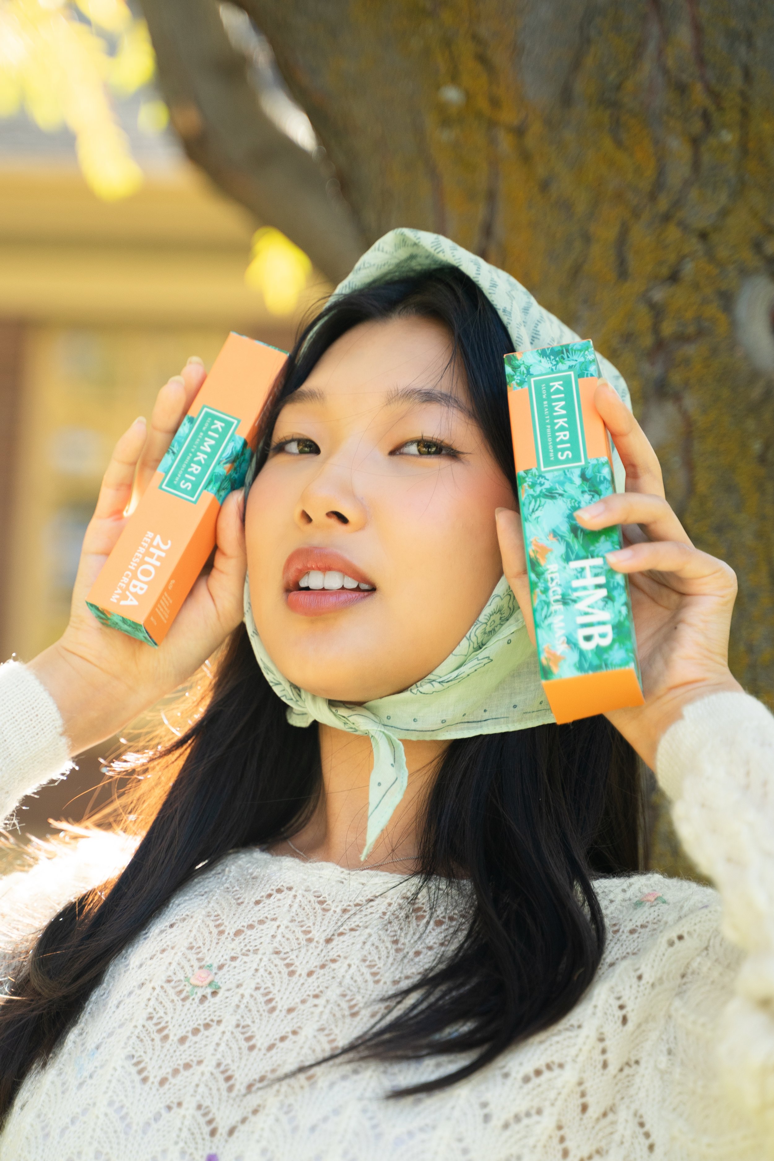 A woman with long black hair, wearing a cream-colored lace top and a patterned headscarf, holding two boxes of Kimkris bath soap near her ears, outdoors with a blurred tree and sunlight in the background.