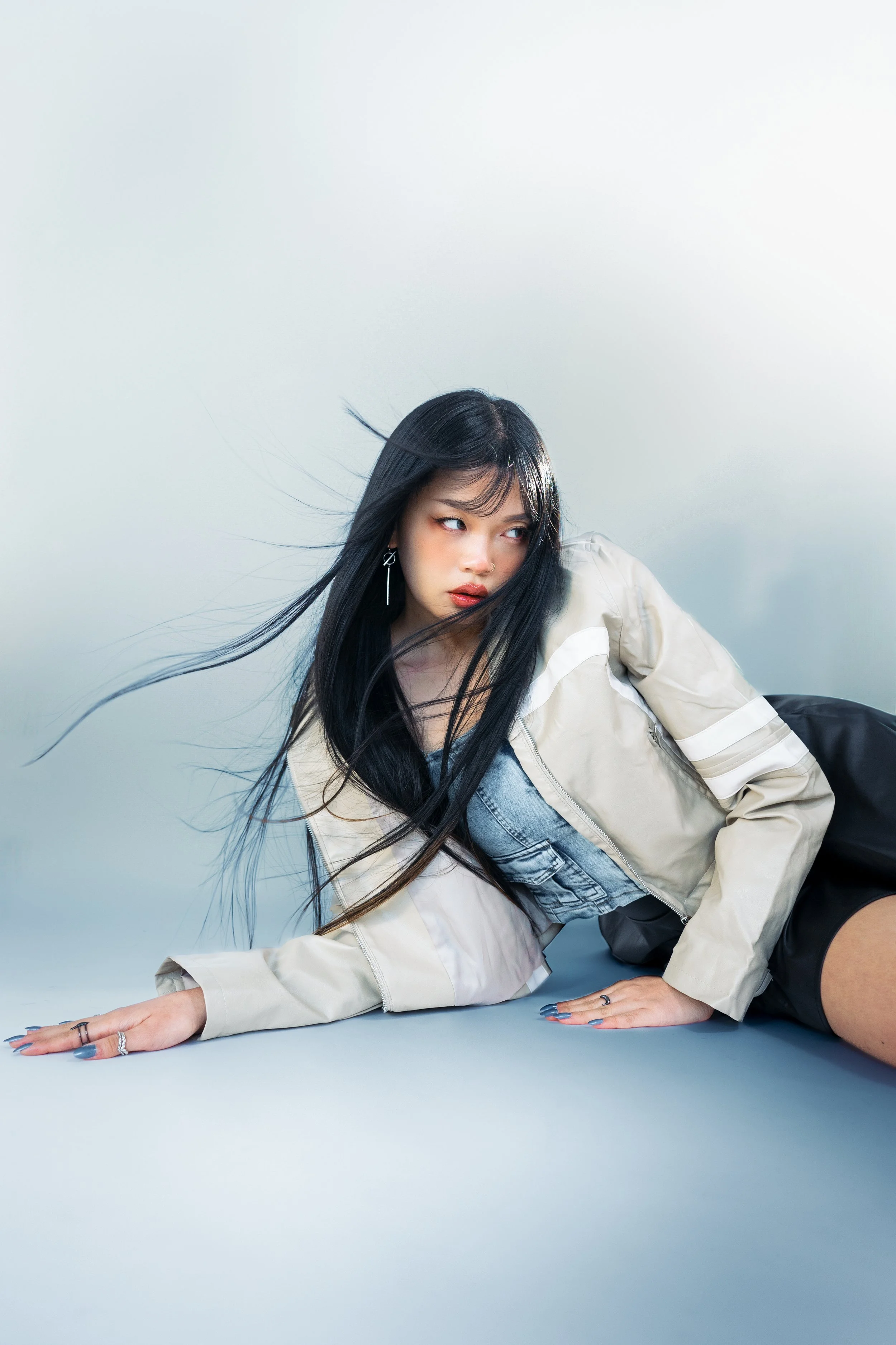 Young woman with long black hair lying on the floor, looking sideways, wearing a beige jacket, denim shirt, black shorts, and multiple rings, with windblown hair and a neutral expression.