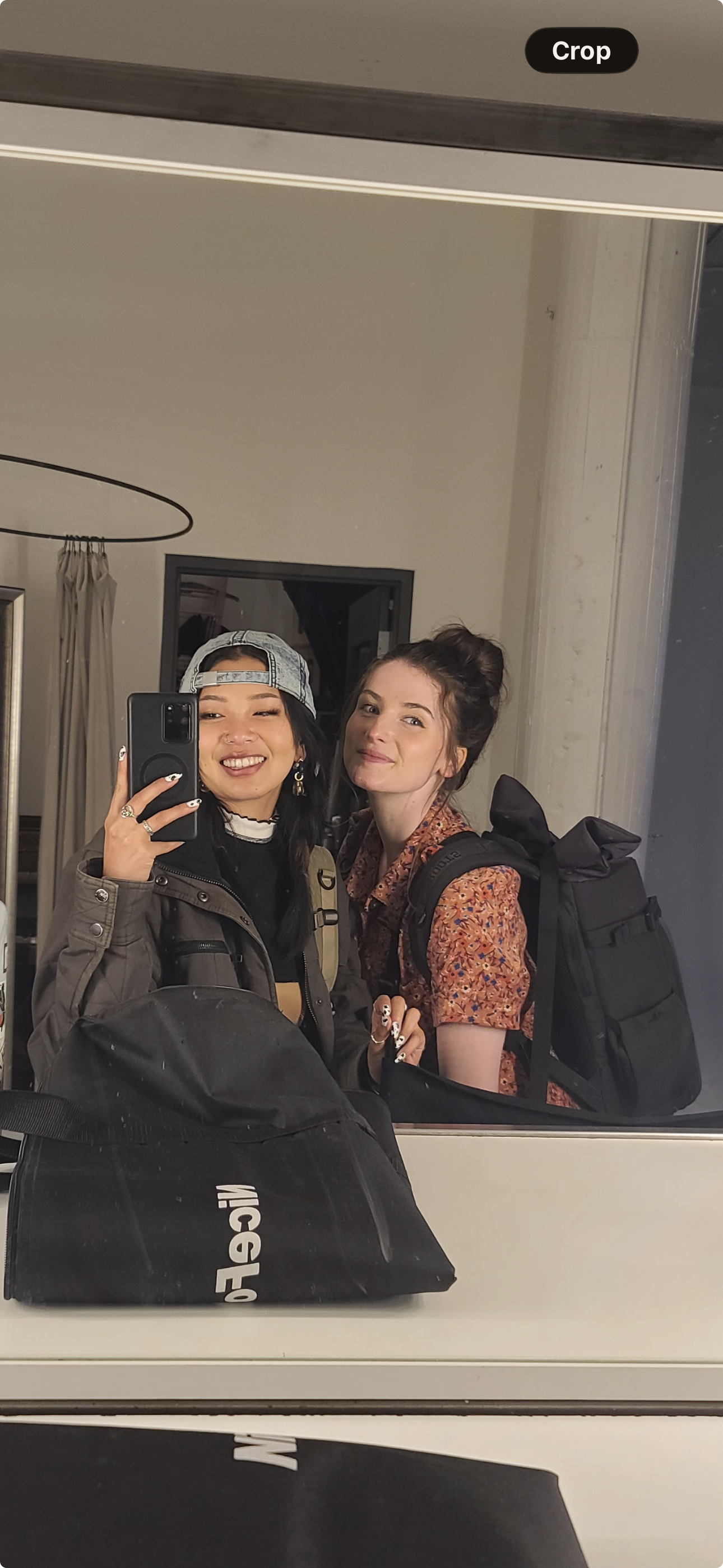 Two young women taking a selfie in front of a mirror, both smiling, with backpacks and casual clothing.
