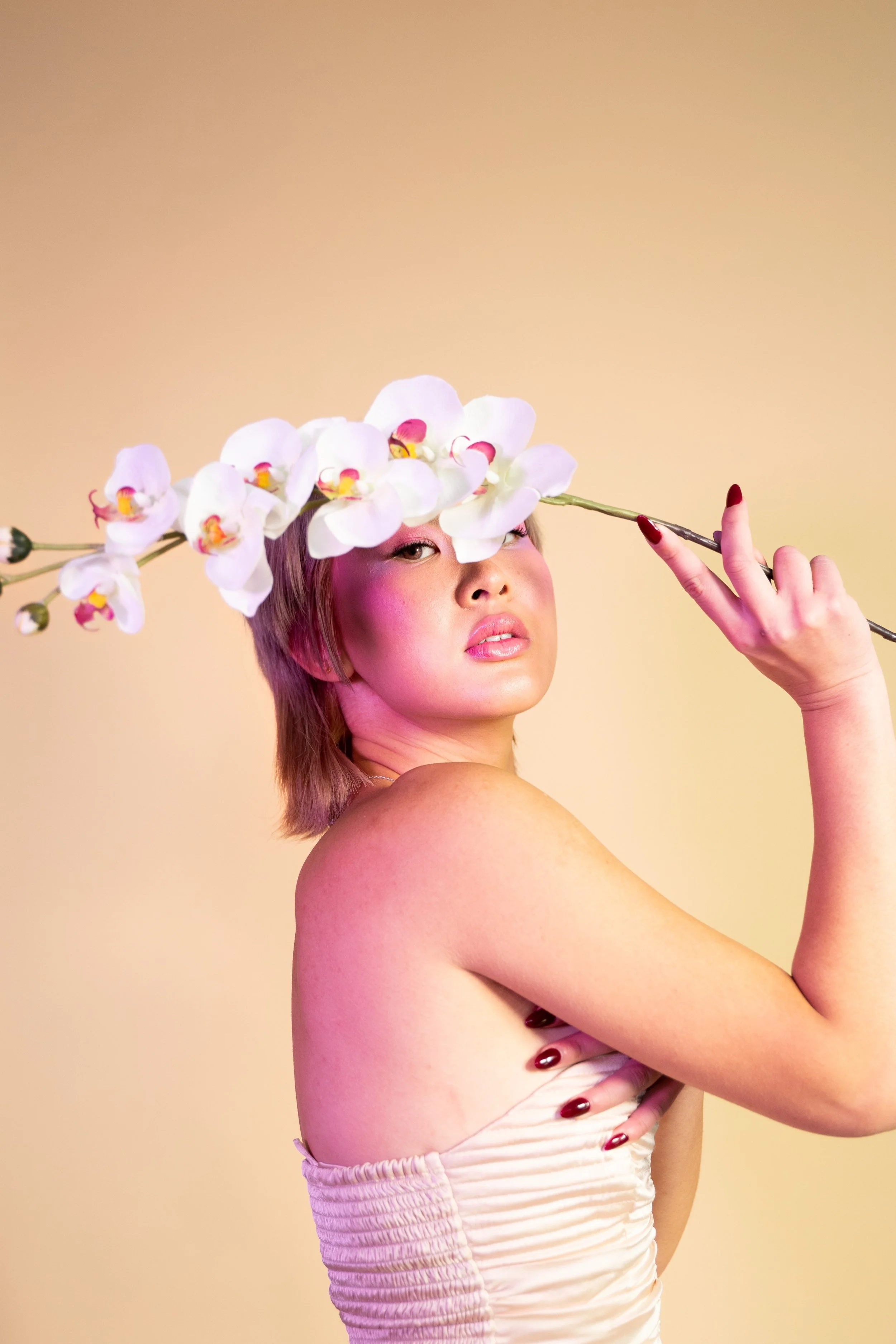 A woman with short blonde hair holding a stem with white orchids, covering her forehead. She is topless, with one arm crossed over her chest, and is wearing a light-colored, ruched fabric garment around her waist. The background is a gradient of beig