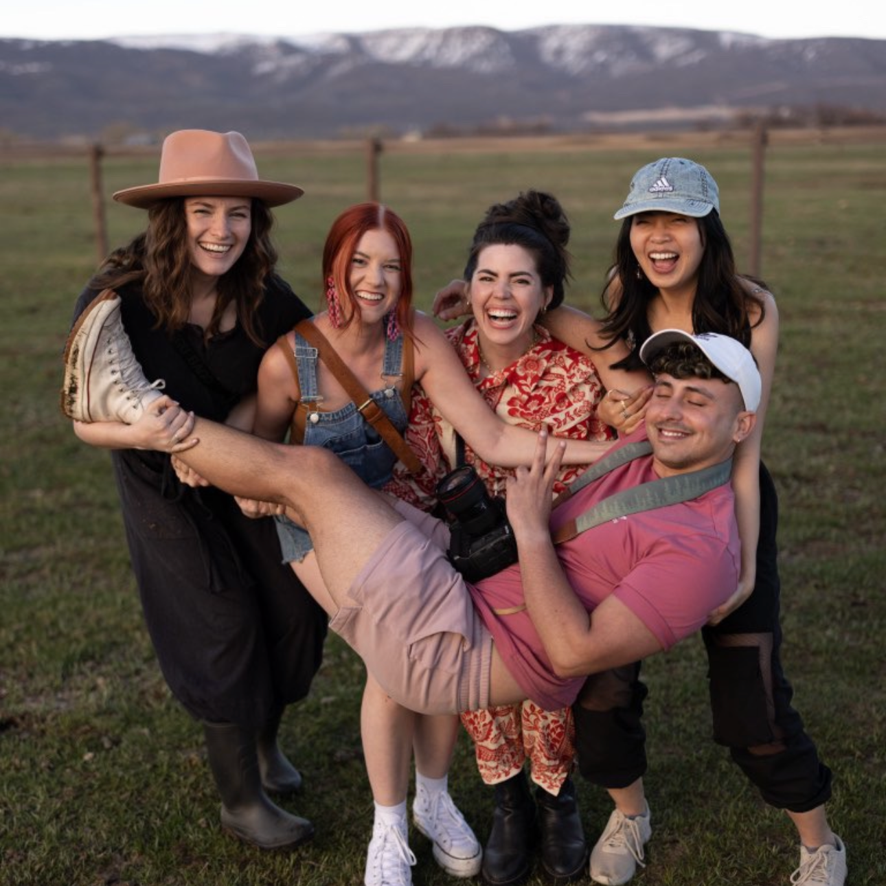 Group of five friends outdoors, three women and one man, laughing and smiling, with mountains in the background, the man being lifted by the women.