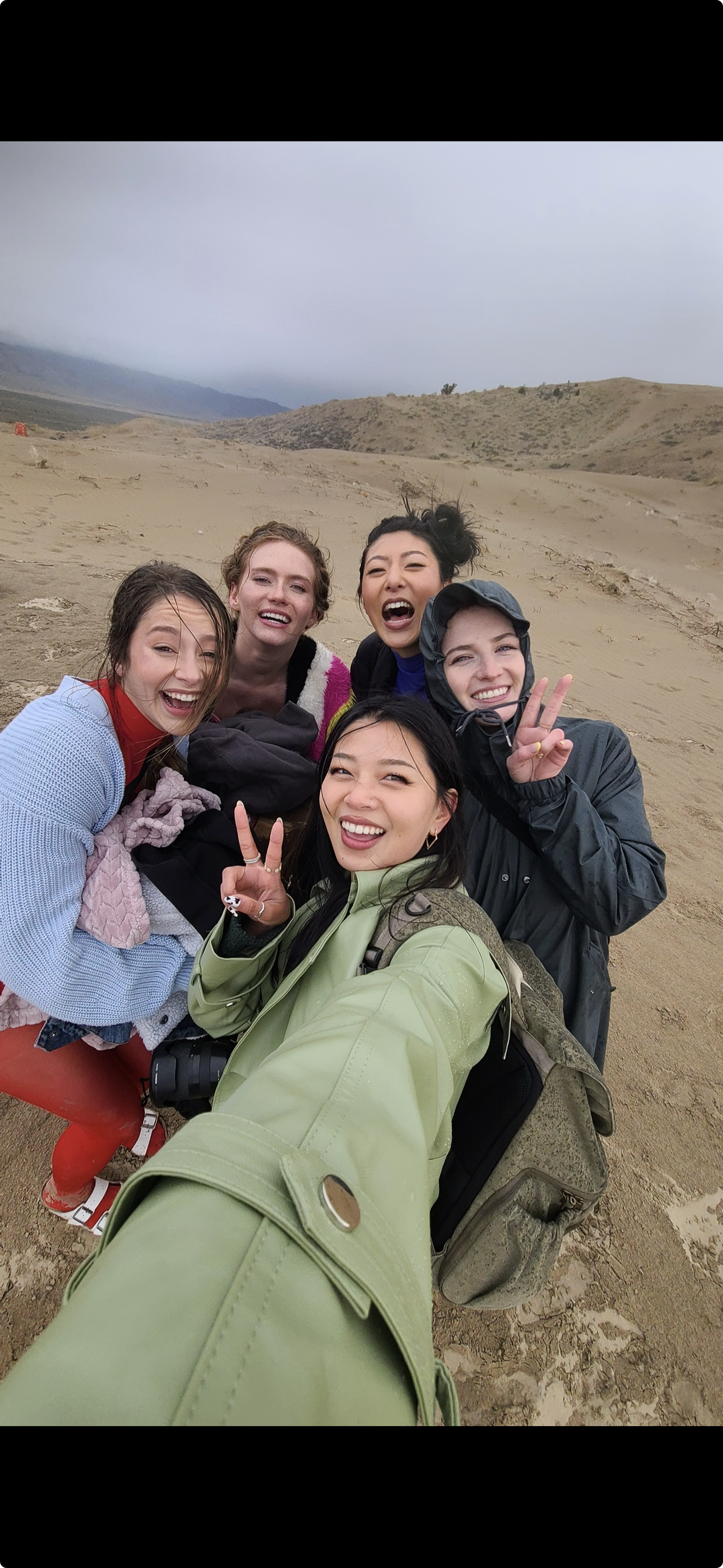 Group of five friends taking a selfie on a sandy, windswept beach on a cloudy day.