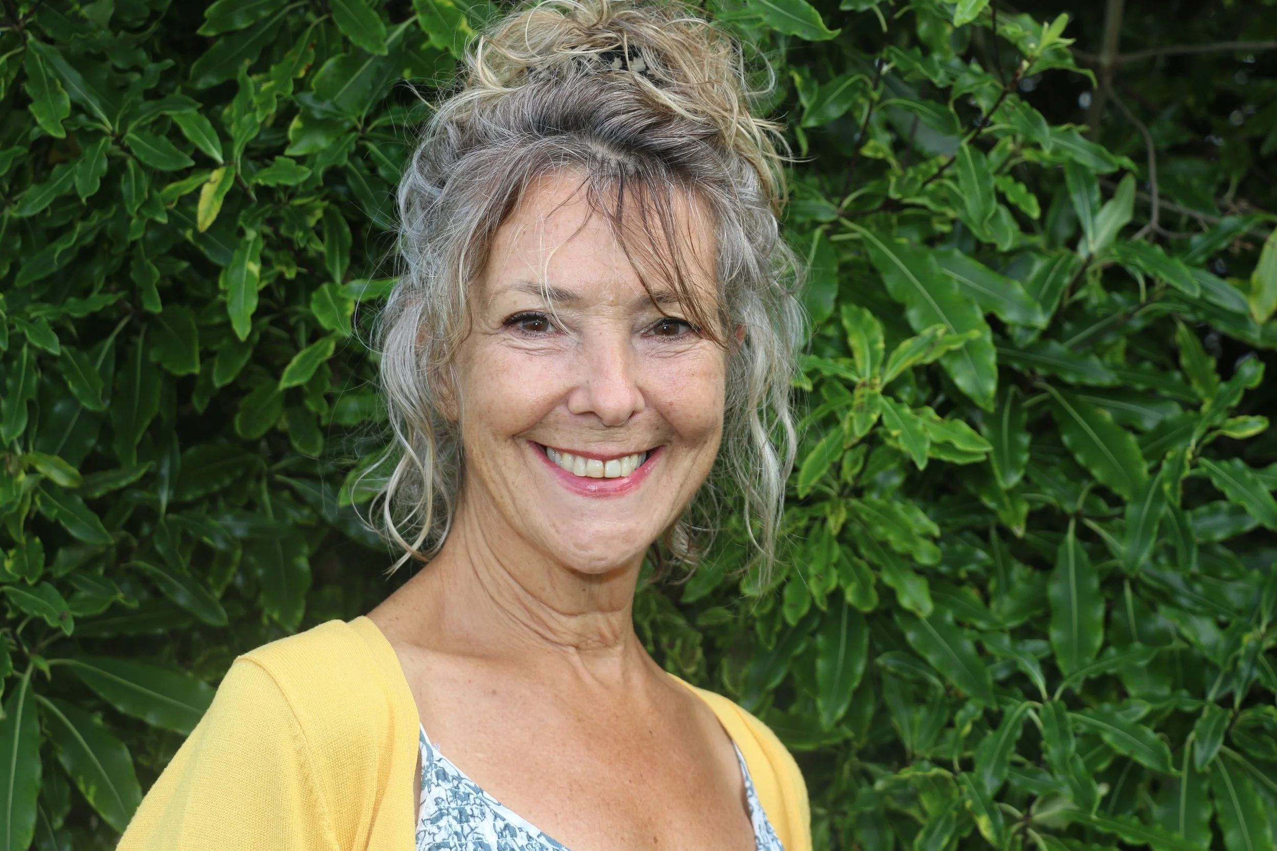 Photo of Service Development Coordinator Kate Smeele. A woman poses for the camera smiling broadly, she is standing in front of a tree with her hands in her pockets wearing a black jumpsuit and grey cardigan. 