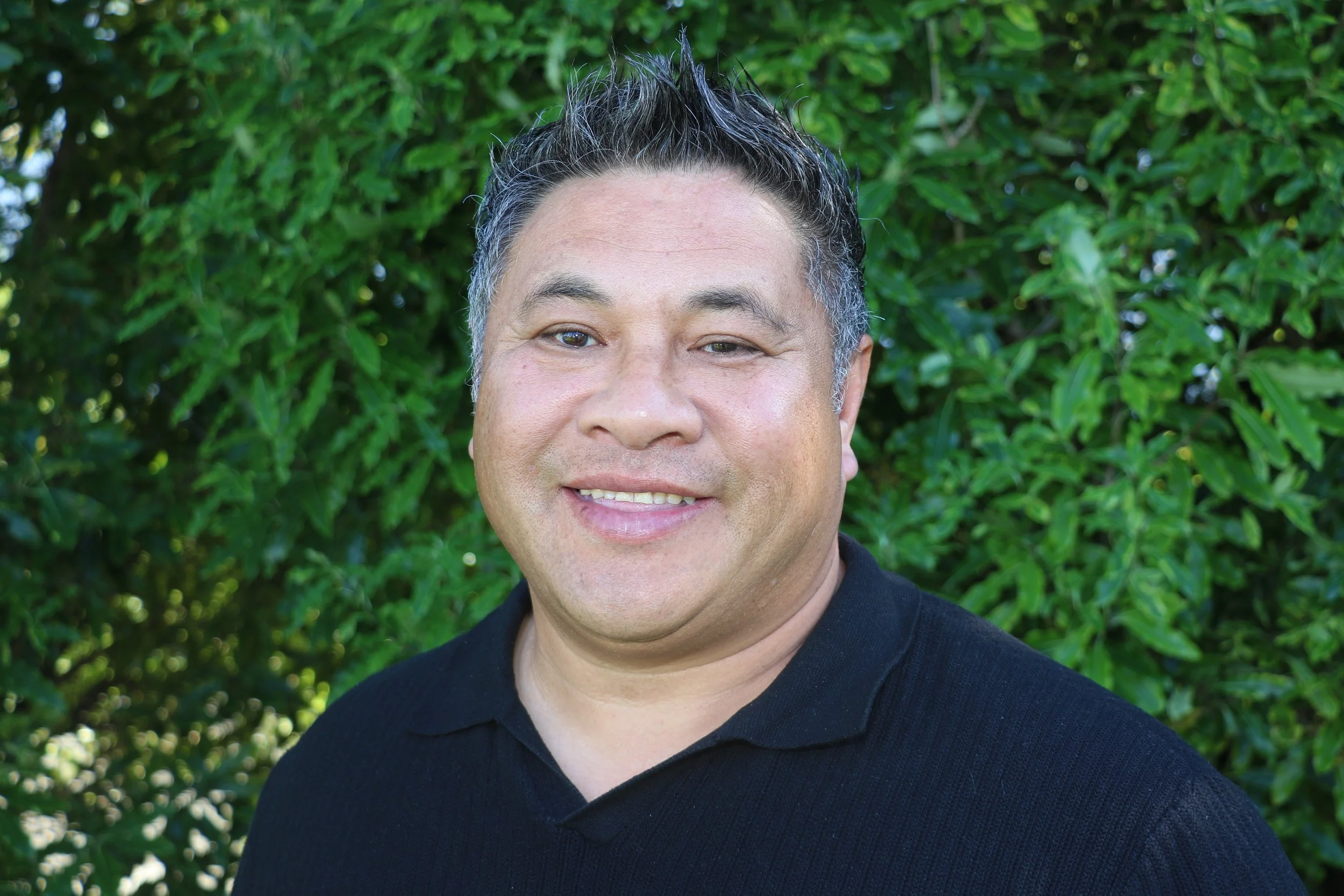 Photo of Service Manager Gafa Tuiloma. A man with black hair poses for the camera standing outdoors in front of a tree. He is wearing a checked shirt tucked into brown trousers. 
