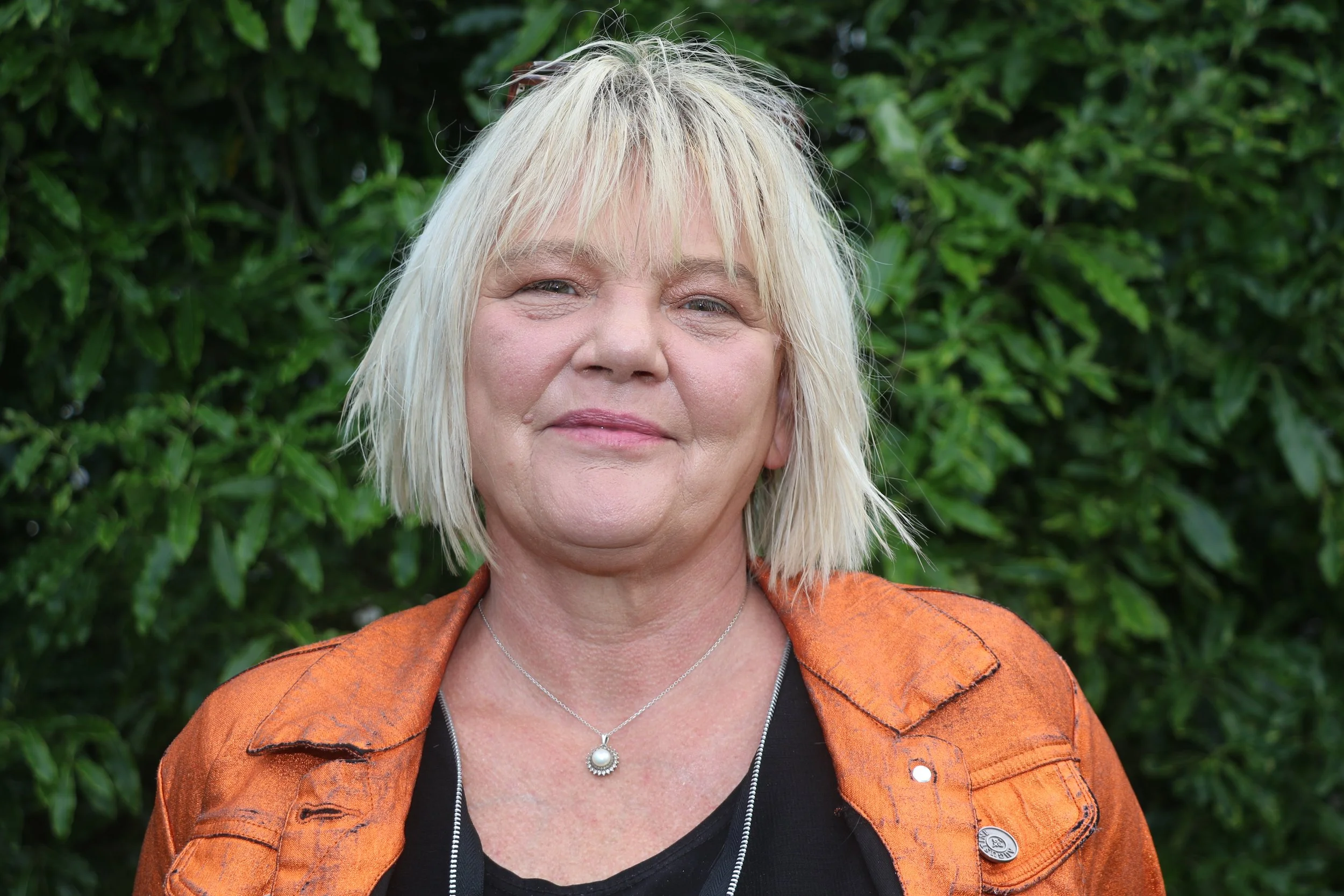 Photo of Operations Manager Vivienne Lorgelly. A smiling woman poses for the camera outdoors, she is standing in front of a tree and is holding a medical mask. She is wearing a grey top and has short white hair.
