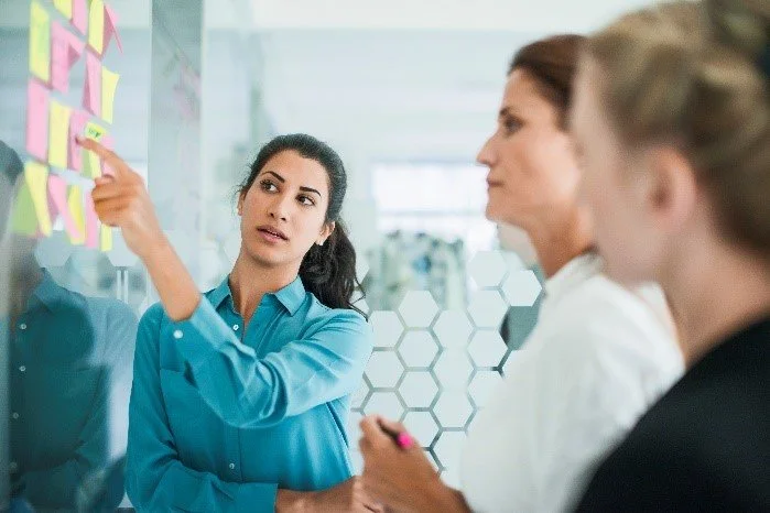 Businesswoman leading a meeting, pointing at sticky notes on a glass wall, with colleagues listening.