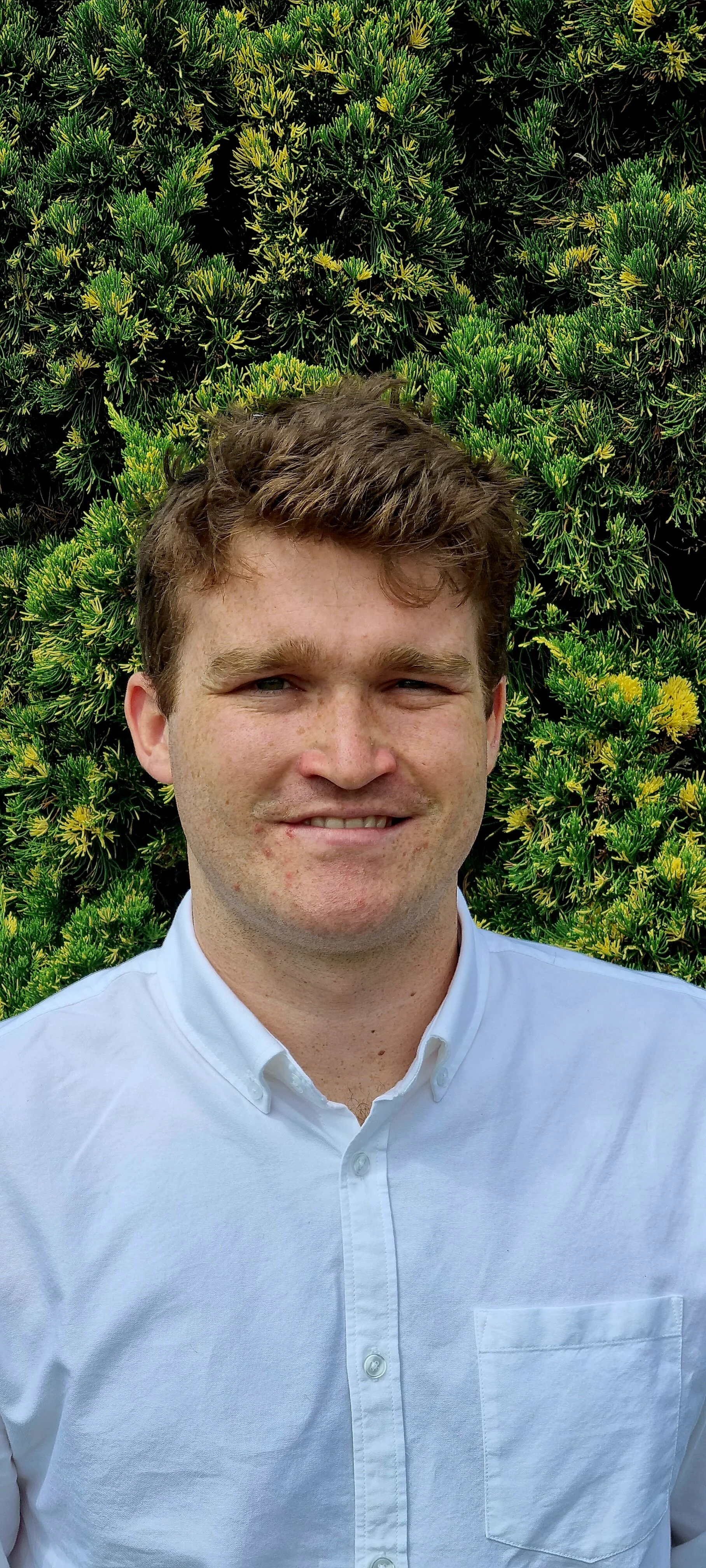 Portrait of a middle-aged man with short light brown hair, wearing a white collared shirt, smiling slightly, standing outdoors against a green leafy hedge background.