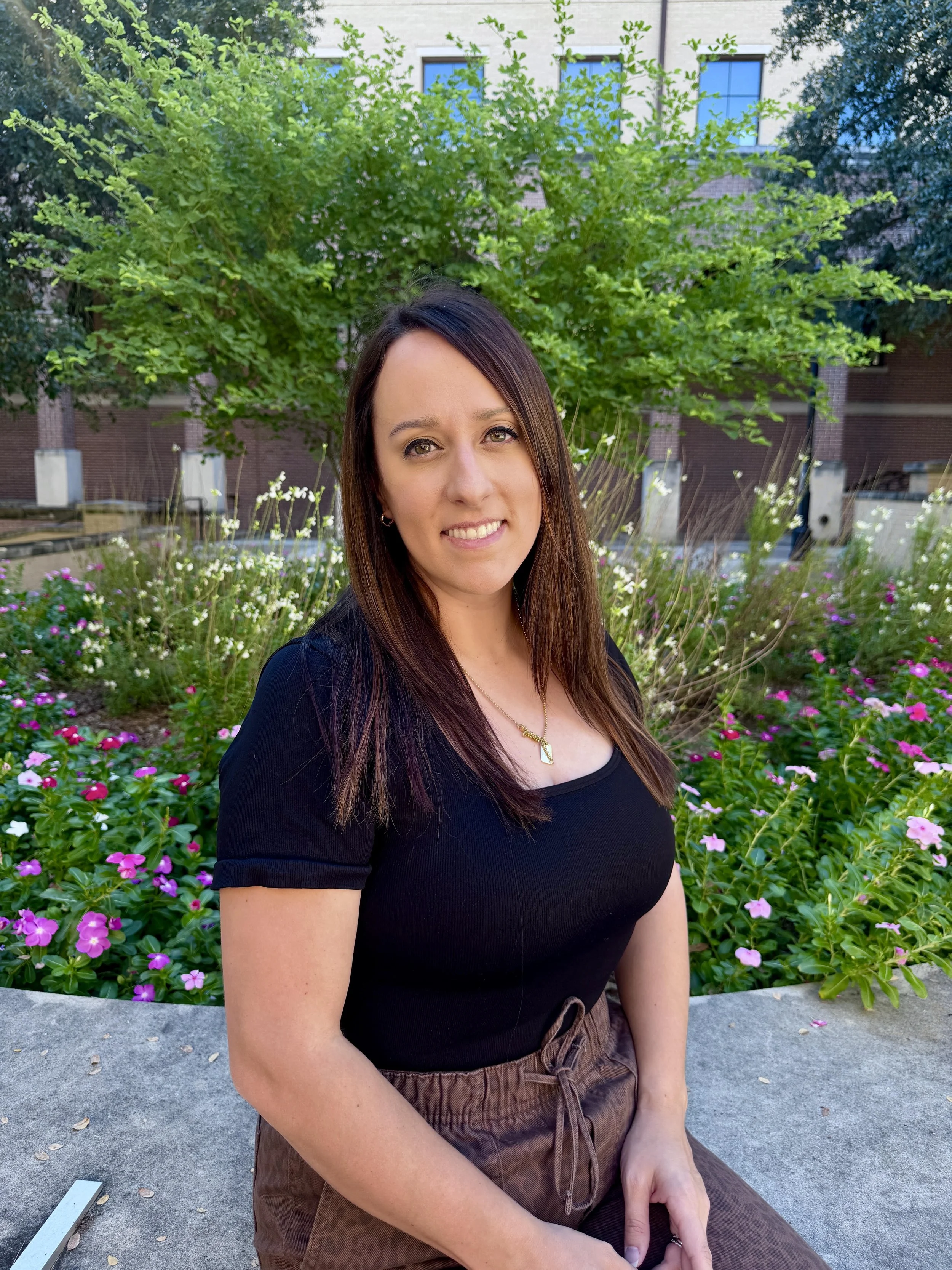 A woman with long, light brown hair standing in front of a brick wall. She is smiling, wearing a black top and a patterned blazer.