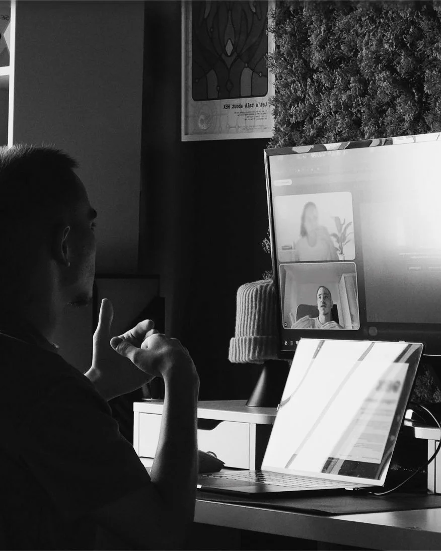 A person engaging in a video call with two women on a computer and a laptop, in a home office setting, in black and white.