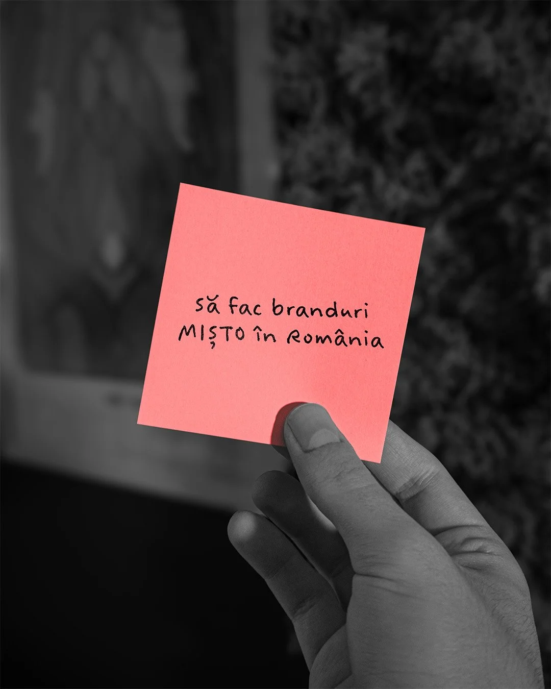 A hand holds a pink sticky note with handwritten text in Romanian, which says 'să fac branduri MISTO în România'.
