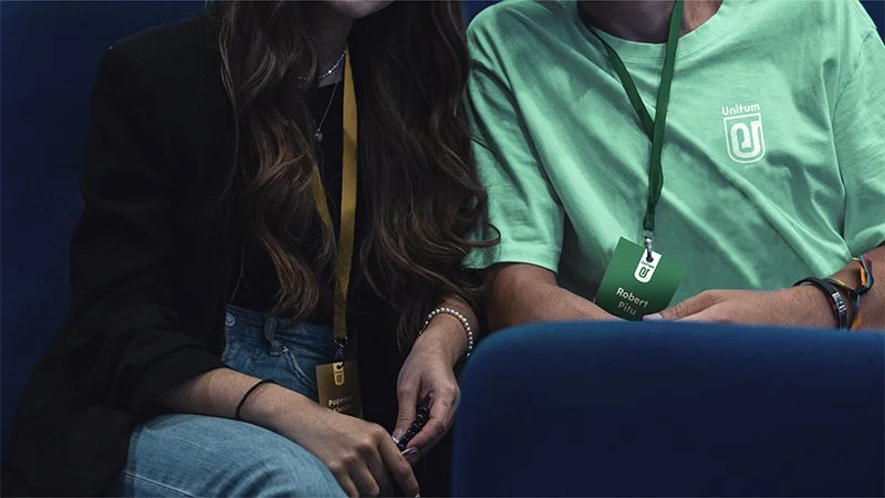 Two people sitting close together, one wearing a black jacket with long wavy hair, the other in a light green T-shirt with a lanyard that says "Robert". They appear to be at an event or conference.