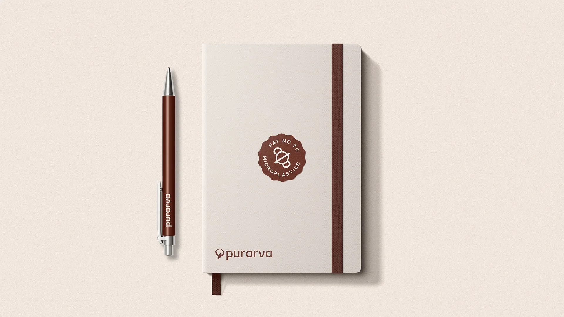 A white notebook with a brown elastic band and a brown logo that says 'Say No To Microplastics' with a small icon, along with a brown and silver pen with the brand name 'Purarva' on it, all placed on a light beige background.