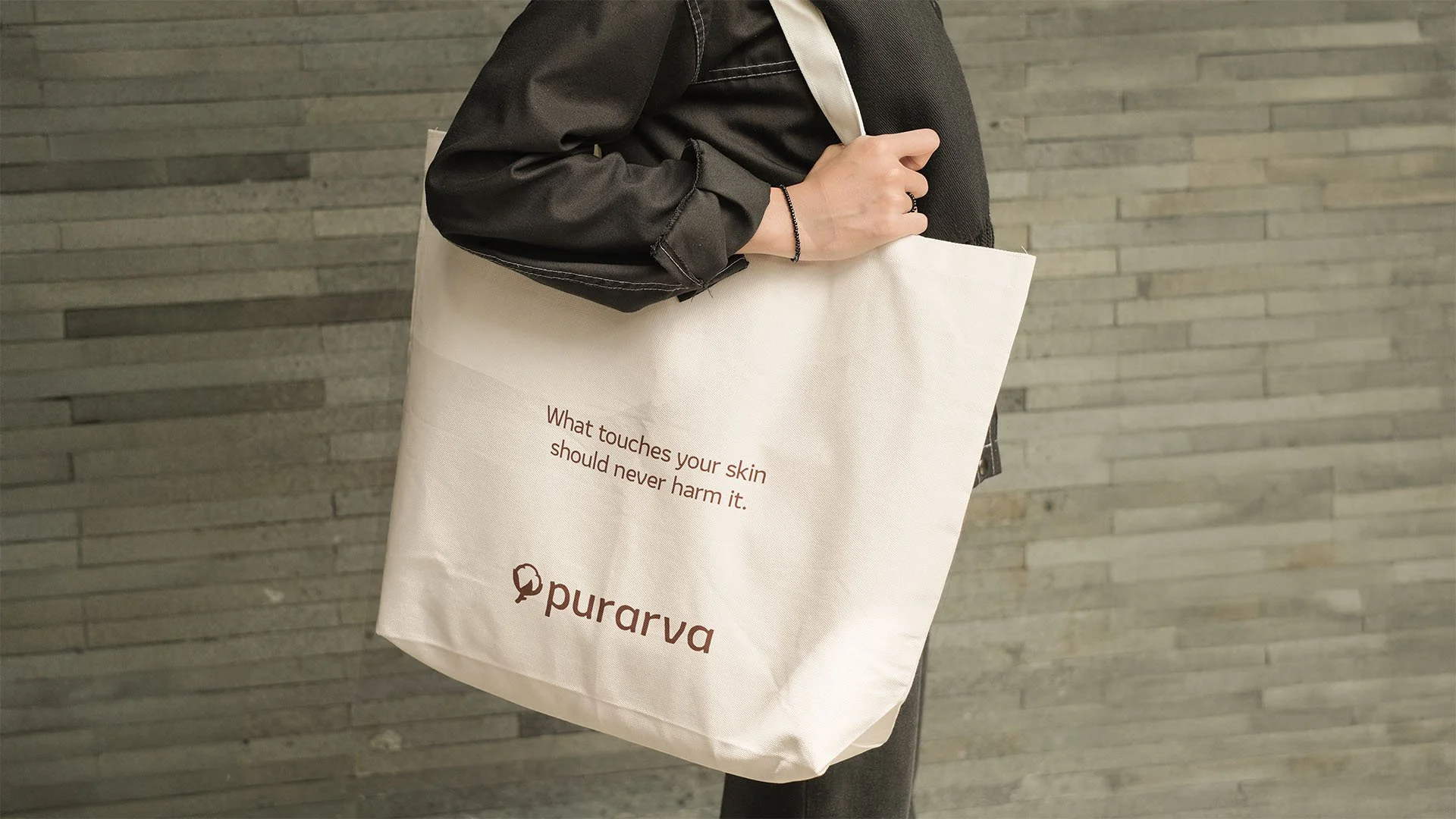 Person holding a white tote bag with a brown logo and text that says, 'What touches your skin should never harm it,' against a gray brick wall background.