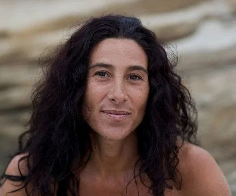 A woman with long, curly dark hair, looking at the camera with a neutral expression, outdoors near rocks or driftwood.