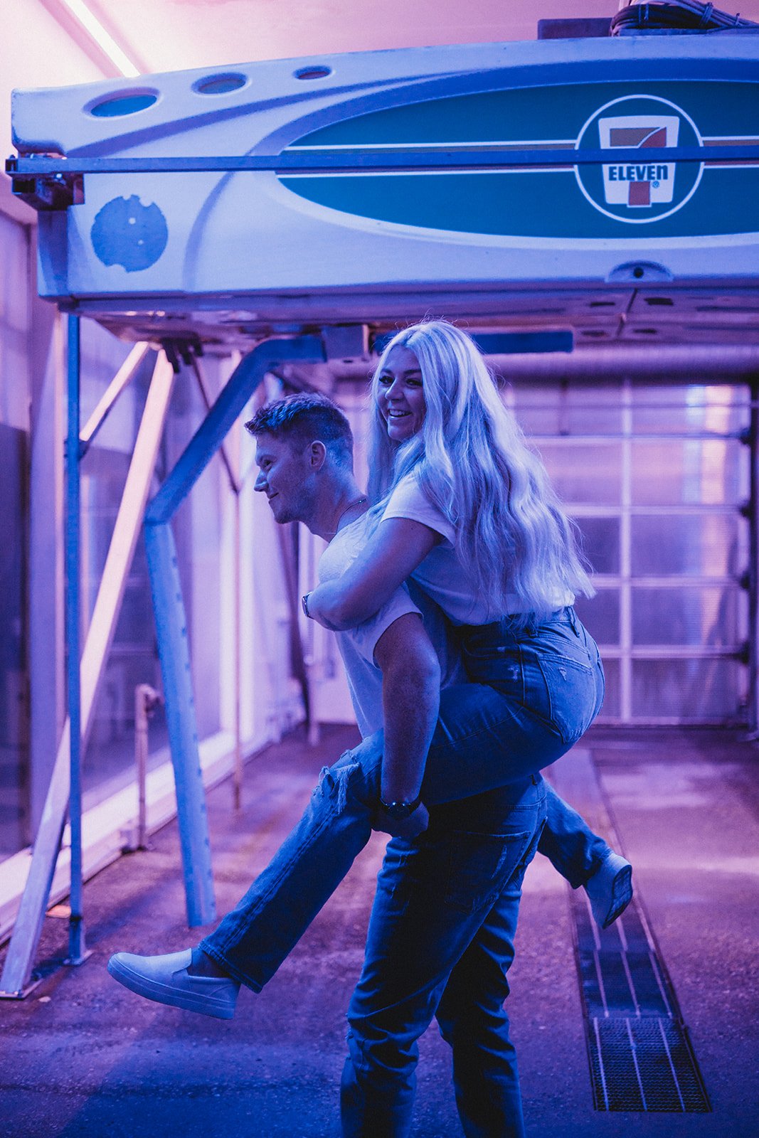 man carrying woman on his back inside of a car wash for their engagement photos, she is laughing as they spin around in Fort Saskatchewan, alberta