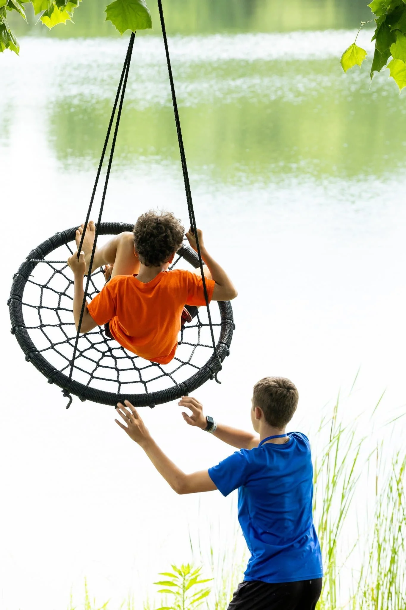 A child rides a round rope swing beside a pond while another person gives a push.