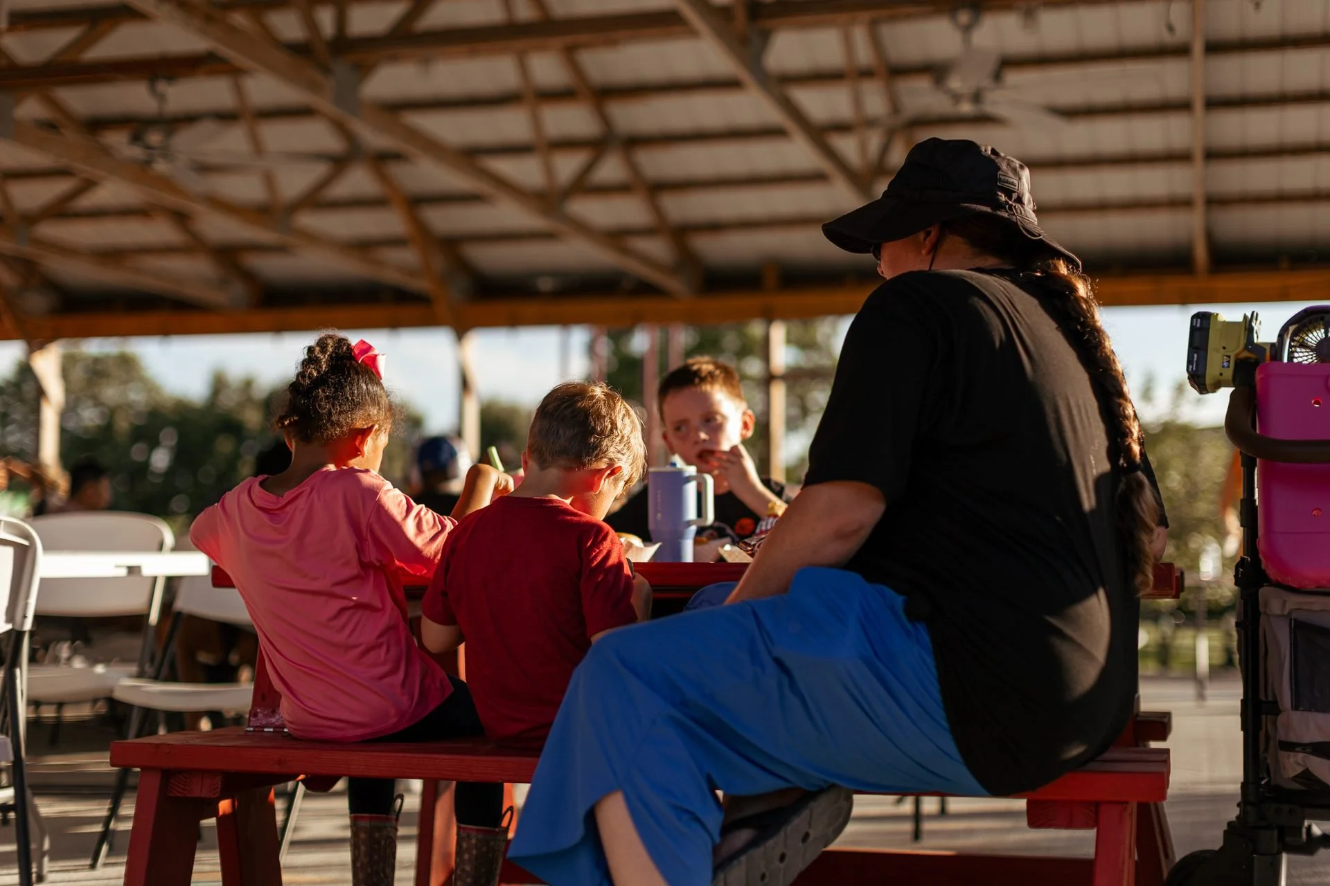 Children and an adult eating together at a red picnic table under an open-air pavilion at Joy Meadows.