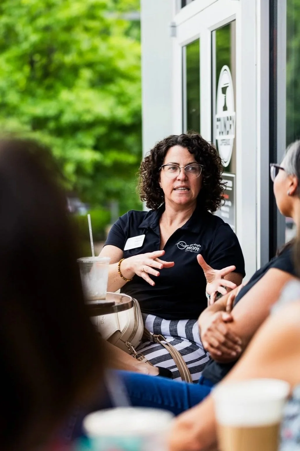 A woman in a Single Mom KC polo speaks with others at an outdoor coffee shop networking event.