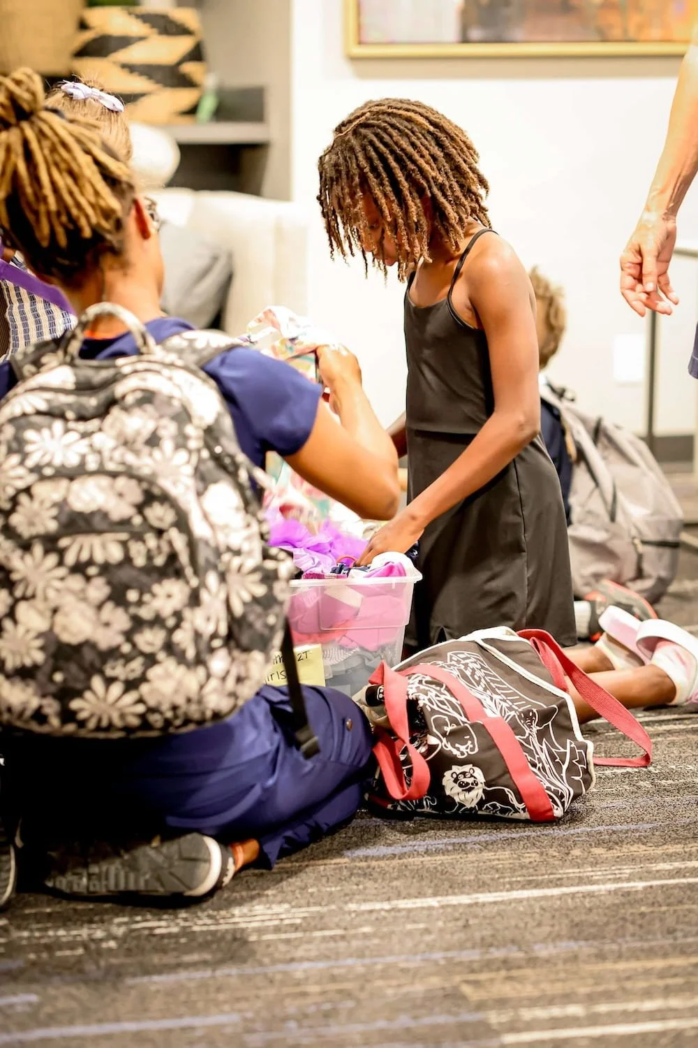 Two girls with backpacks kneel, looking through a plastic bin of resources for children in foster care in a brightly lit room.