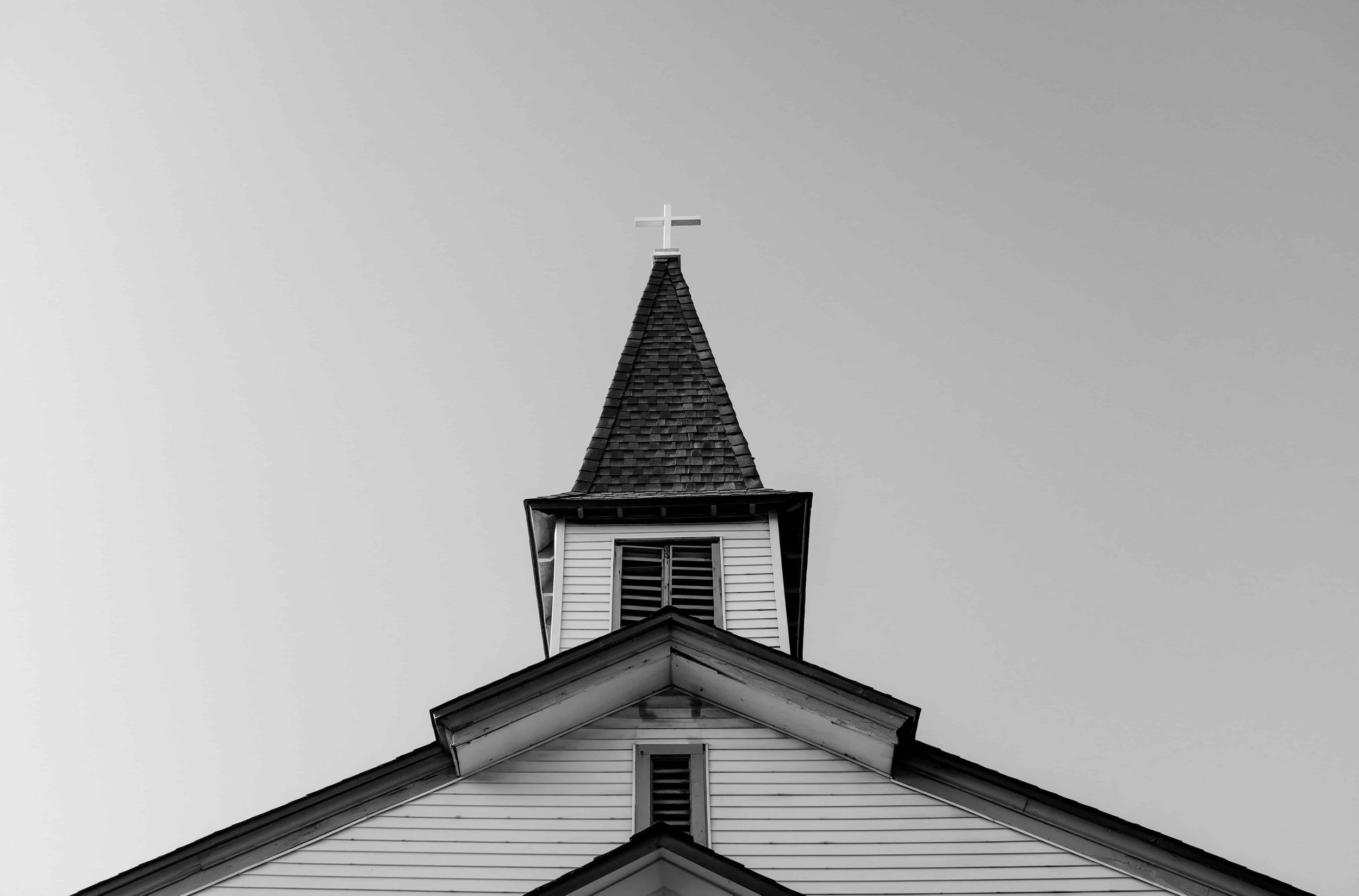 Black and white image of a church steeple with cross against a clear sky, symbolizing faith, community support, and local outreach programs