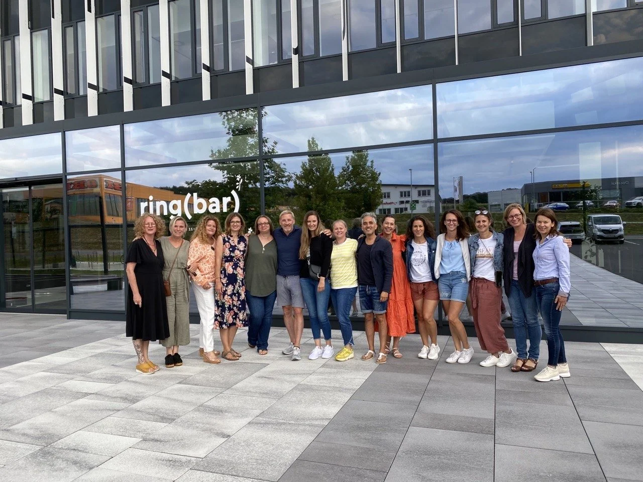 Eine Gruppe von zwölf Personen steht lächelnd vor einem Gebäude mit großen Fenstern. Im Hintergrund ist der Schriftzug "ring(bar)" zu sehen. Alle tragen legere Kleidung und posieren für die Kamera. Vor dem Gebäude sieht man einen gepflasterten Gehweg.