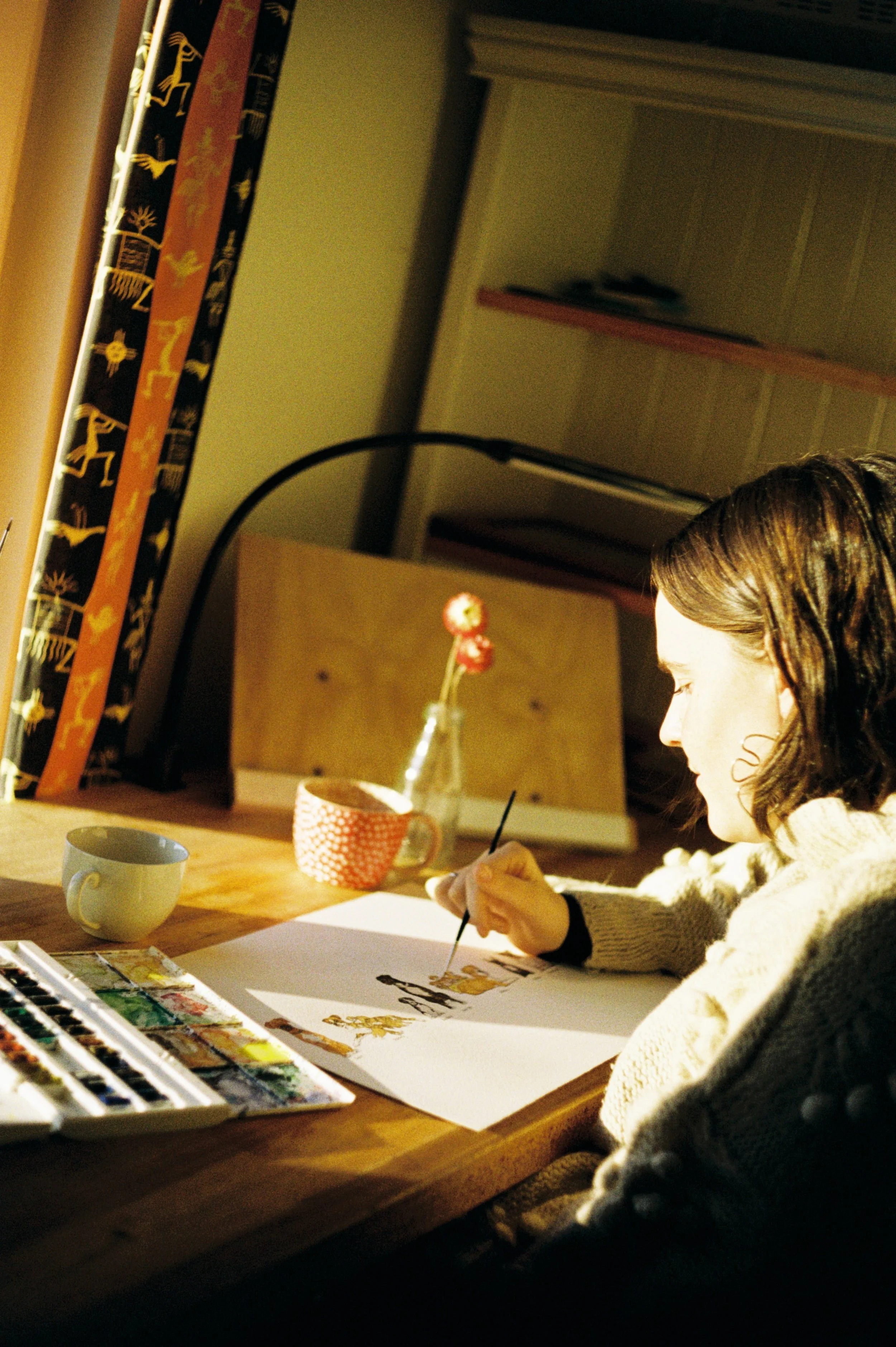 A woman sitting at a wooden desk painting with watercolor paints, with a cup and a vase with two red flowers on the desk.