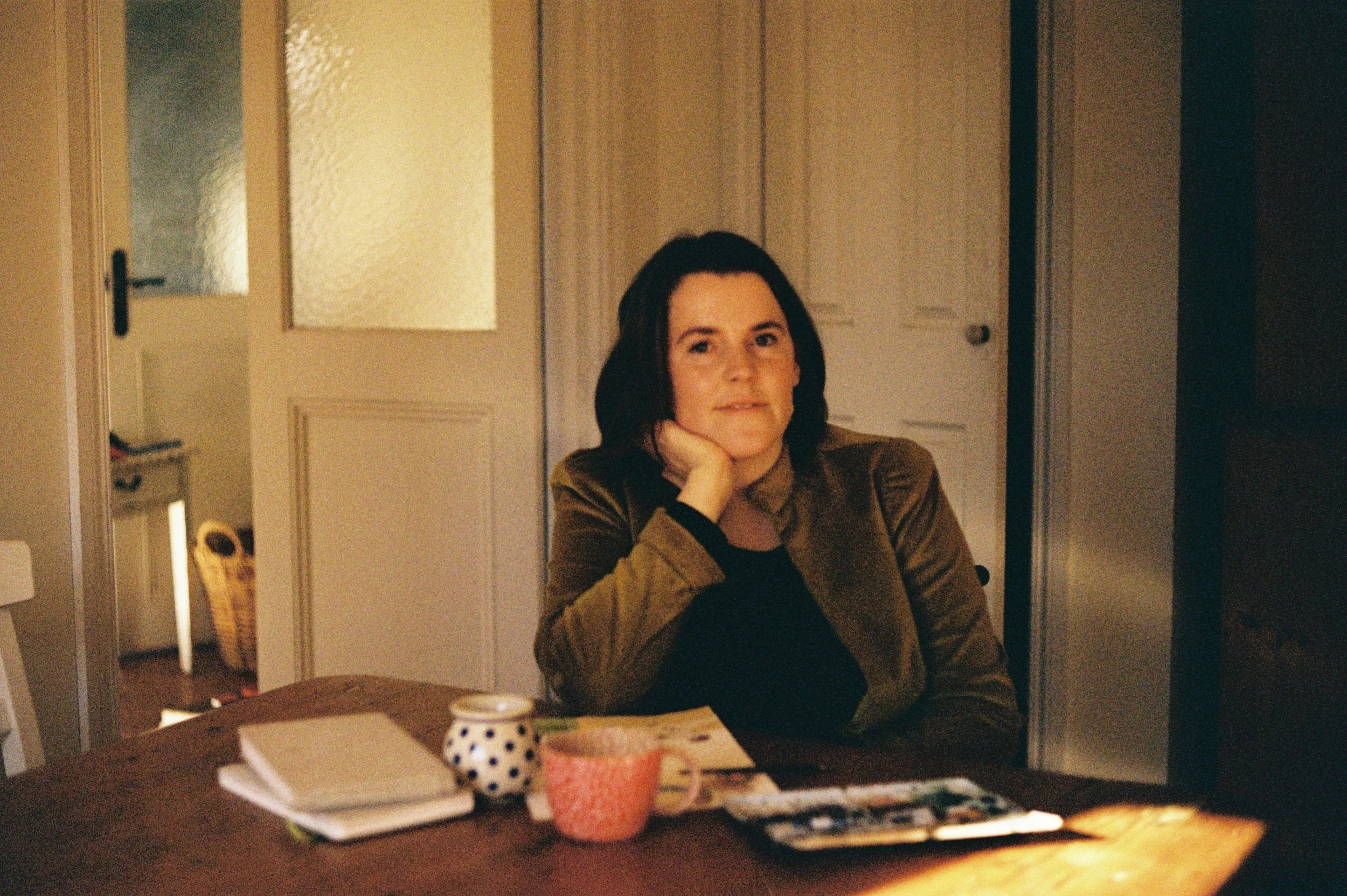 A woman with dark hair sitting at a table in a dimly lit room, resting her chin on her hand, with notebooks, a mug, and a tray of photographs or papers in front of her.