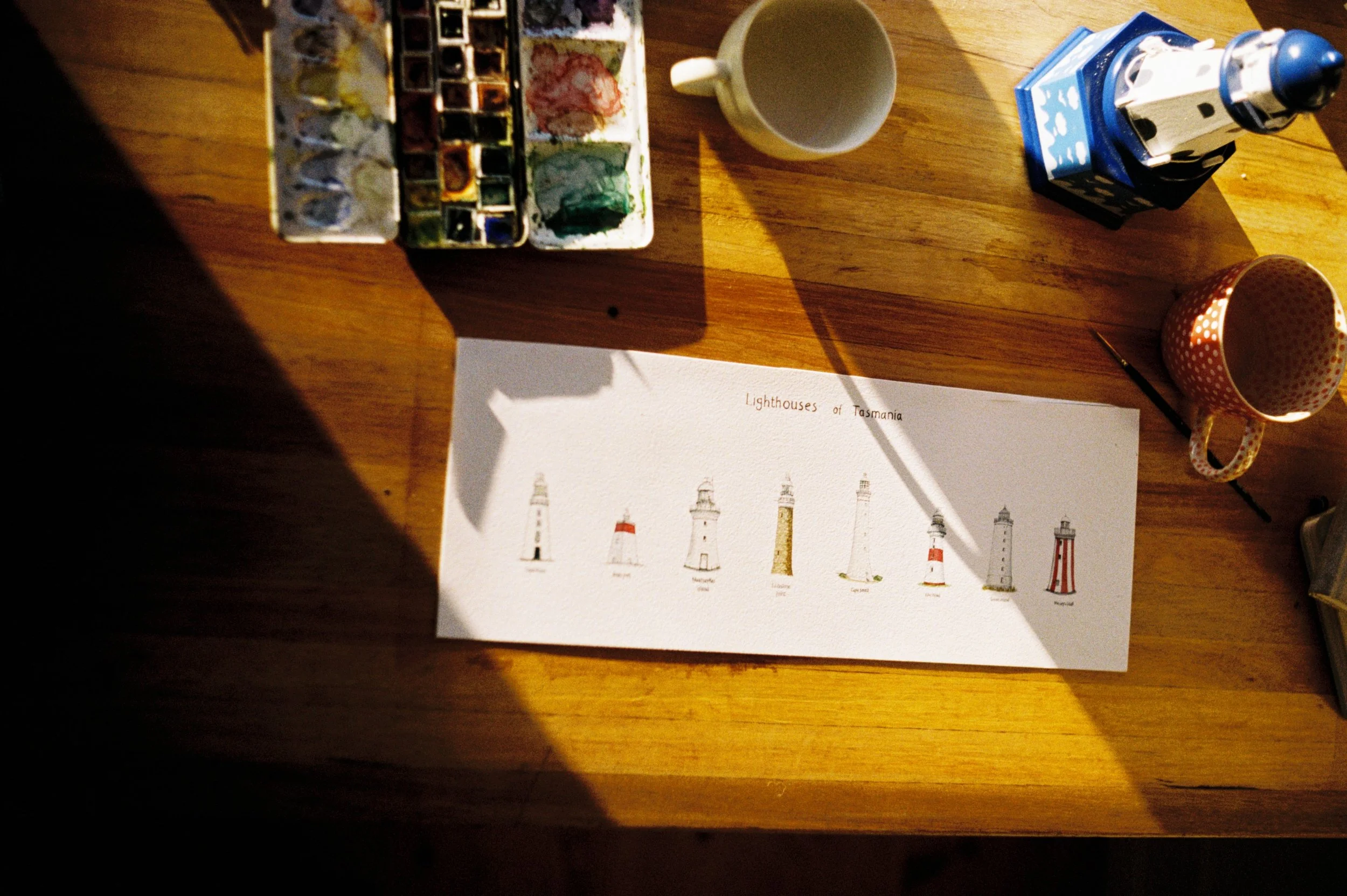 A watercolor painting of different lighthouses of Tasmania on a white sheet of paper on a wooden table, surrounded by watercolor paints, a white mug, a blue and white lighthouse figurine, and a polka-dotted mug.