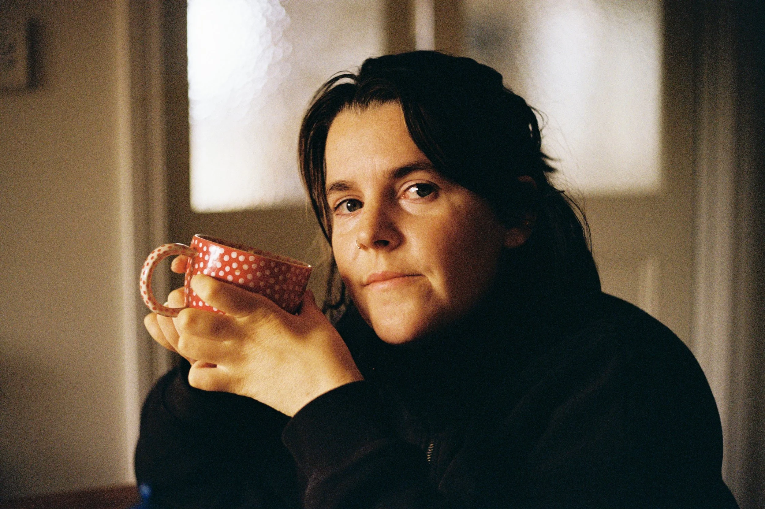 A woman with dark hair and a nose ring holding a red polka-dotted mug, looking at the camera in a cozy indoor setting. Disabled artist, Tasmanian illustrator, mother artist.