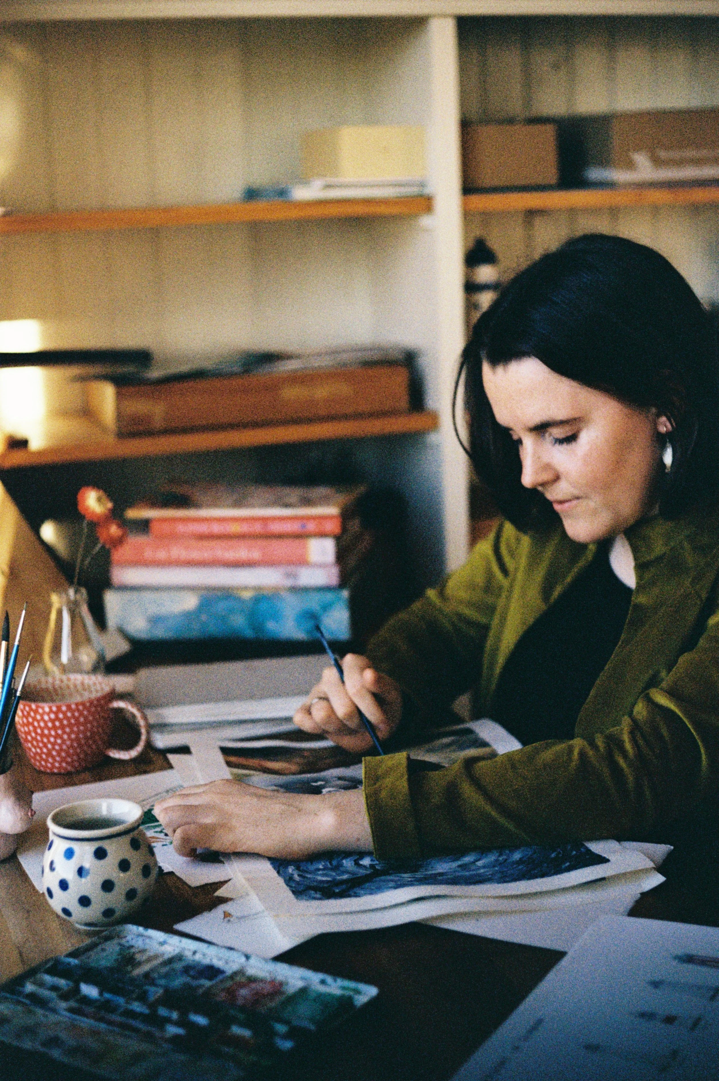 Woman with dark hair, wearing an olive-green jacket, working with paint and papers on a cluttered desk, surrounded by art supplies and books.