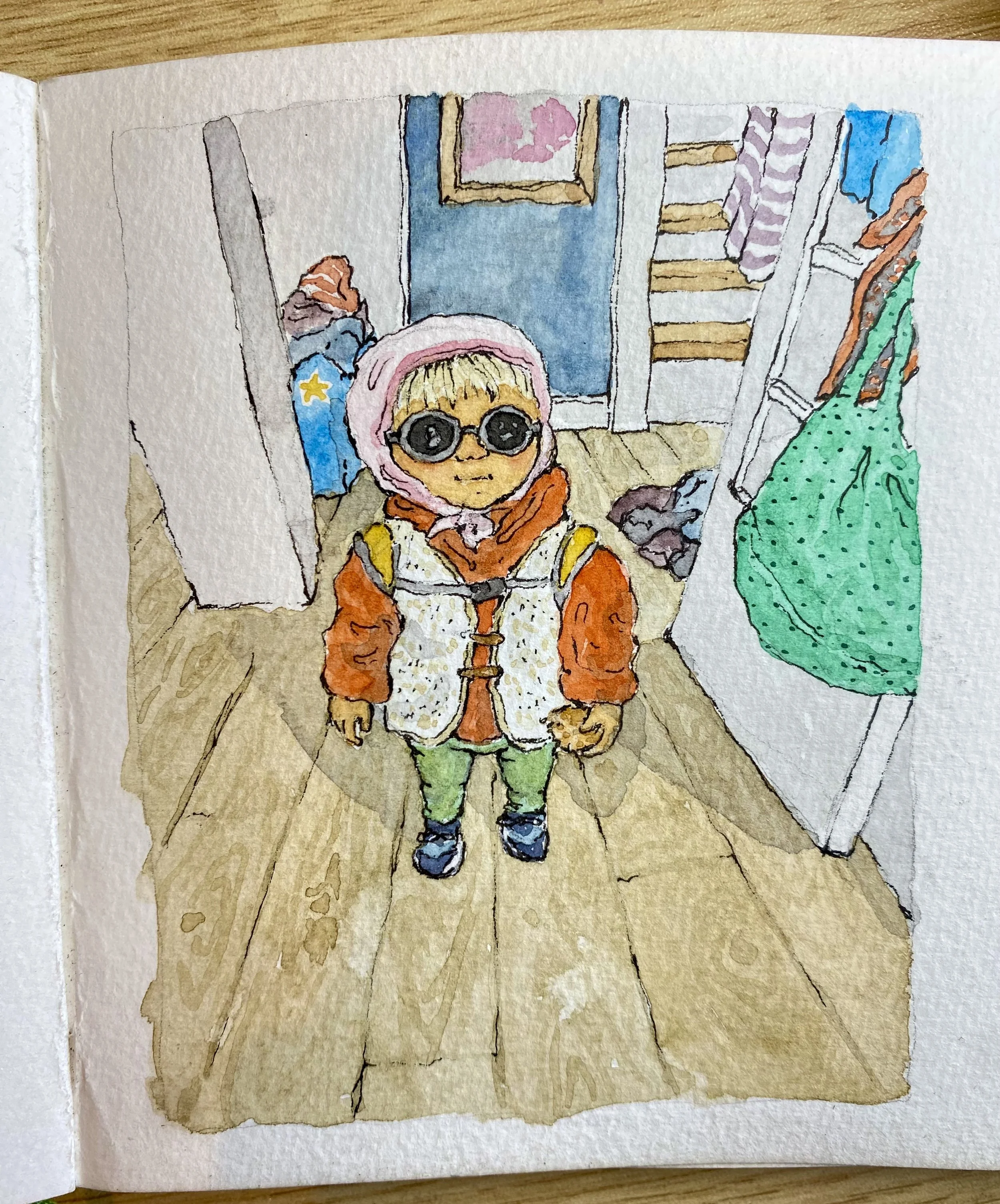 Child wearing sunglasses, pink hood, orange jacket, green pants, and sneakers, standing inside a house with wooden flooring and a staircase surrounded by hanging clothes.