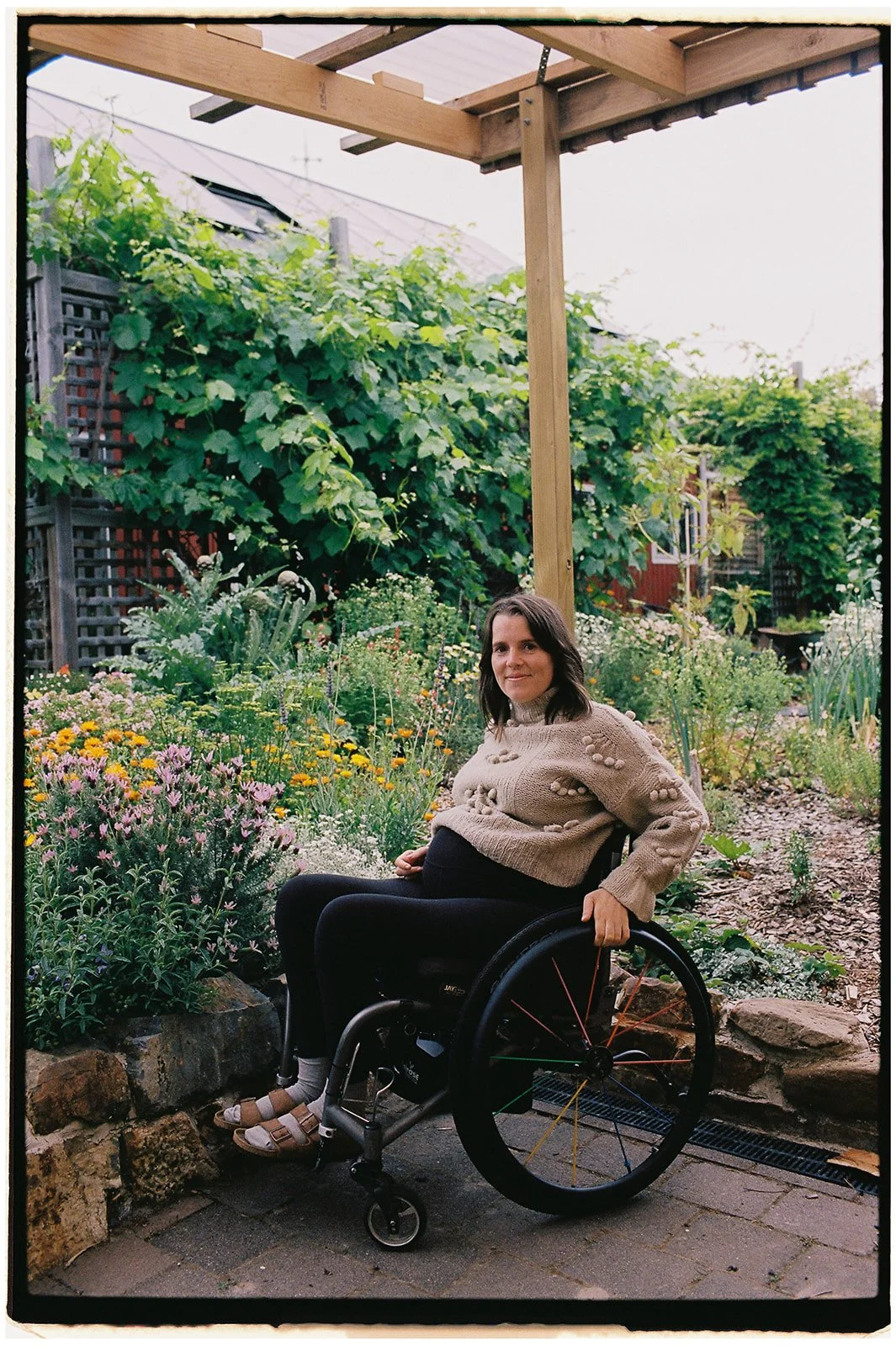 A woman in a wheelchair sitting outdoors in a garden with colorful flowers and greenery, under a wooden structure.
