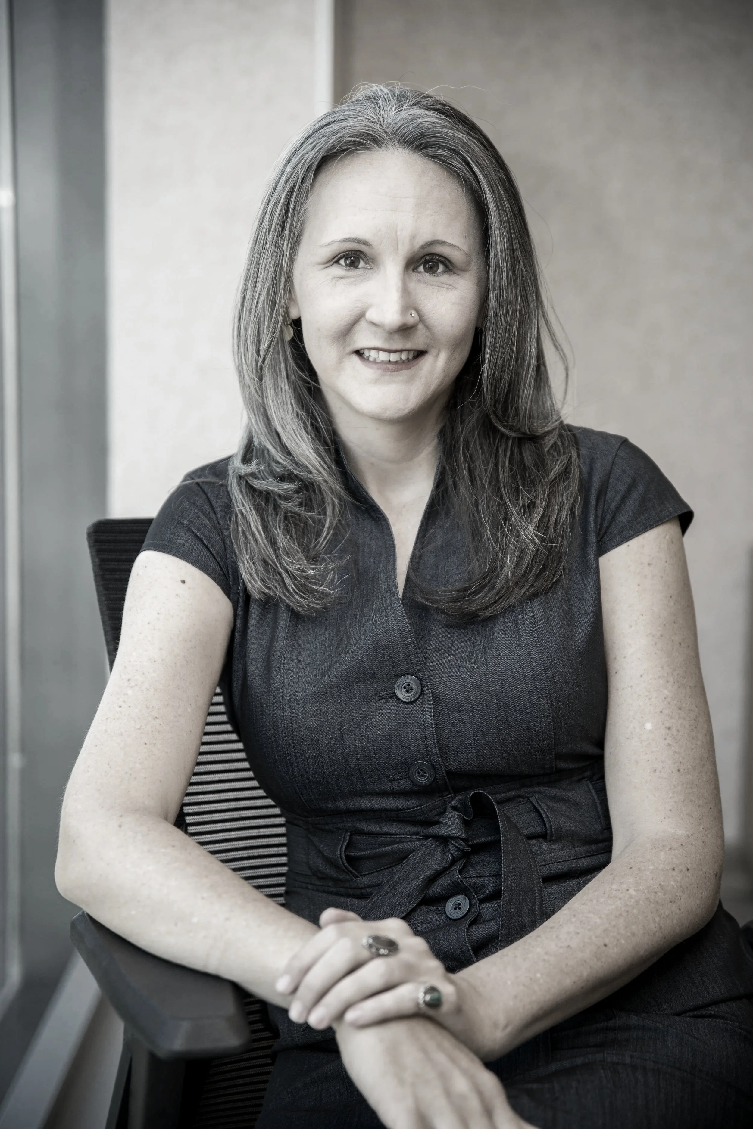 A woman with dark brown hair wearing a pink floral blouse, sitting at a desk with a book, a mug, and a black office chair, smiling.