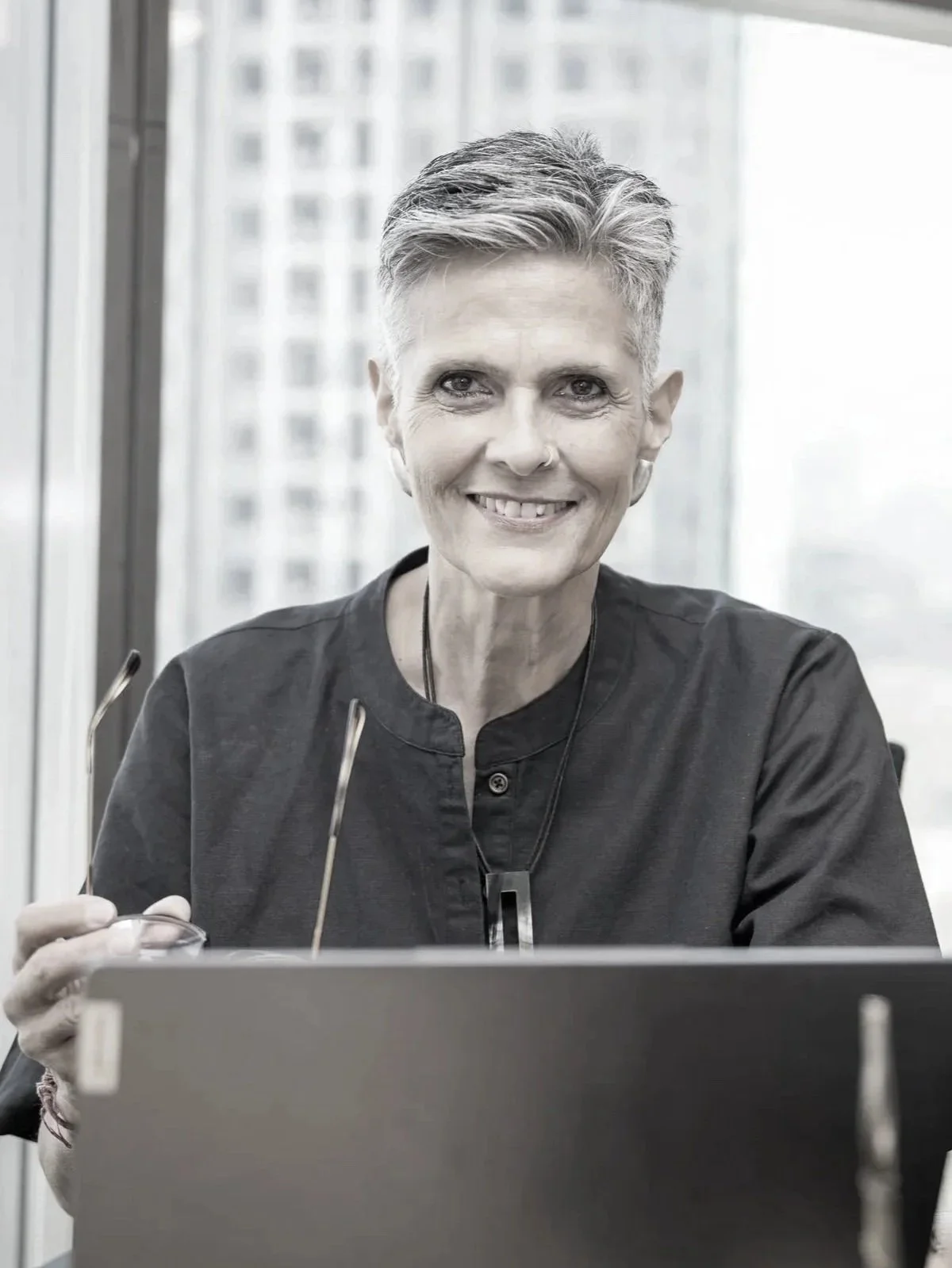 A mature woman with gray hair and glasses, wearing a patterned dress, pearl necklace, and pearl earrings, sits at a desk with her hands clasped, looking confidently at the camera.