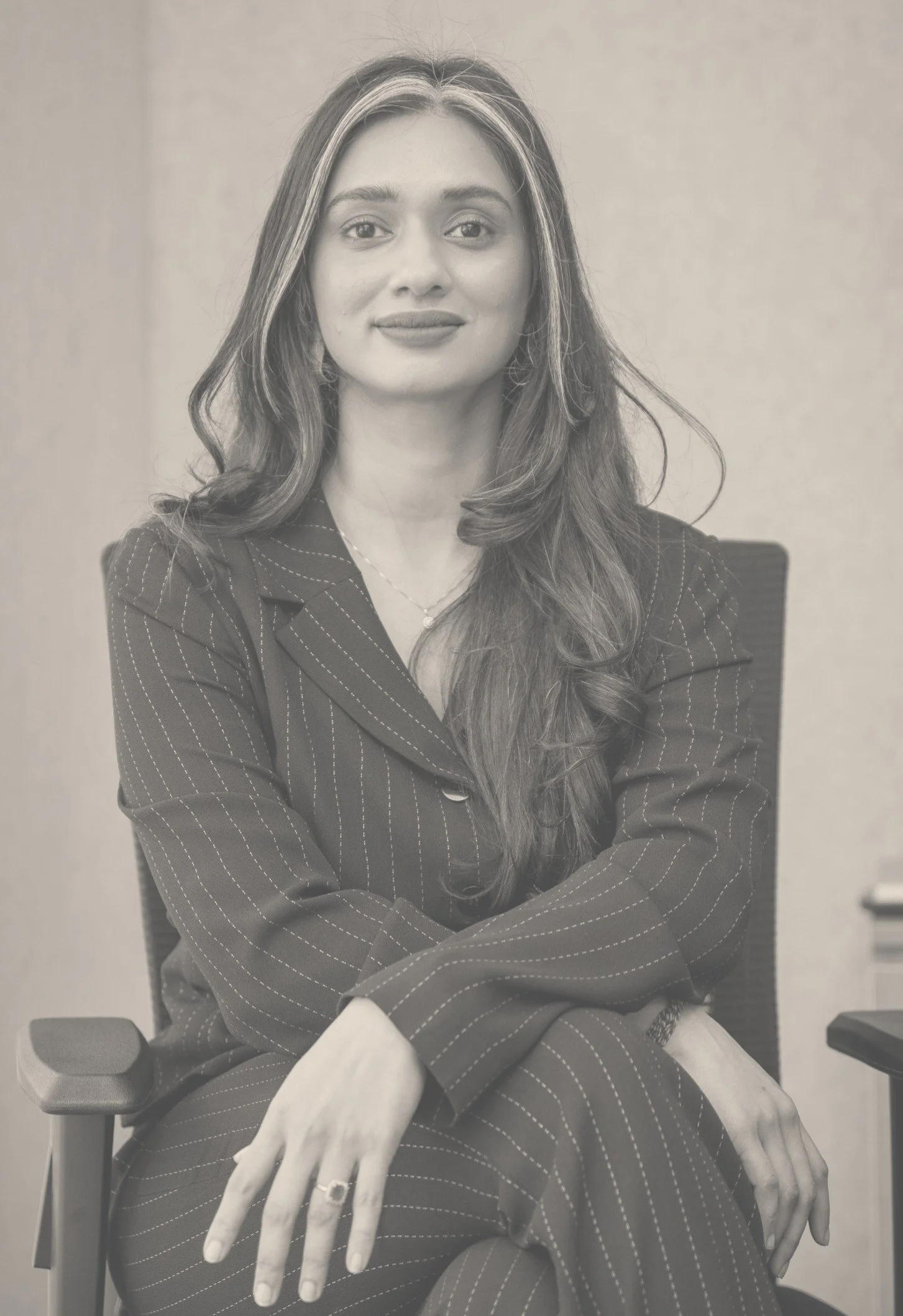 A young woman with long dark hair, wearing a yellow top, sitting at a desk, smiling, with a hand holding a pen near an open notebook in front of her.